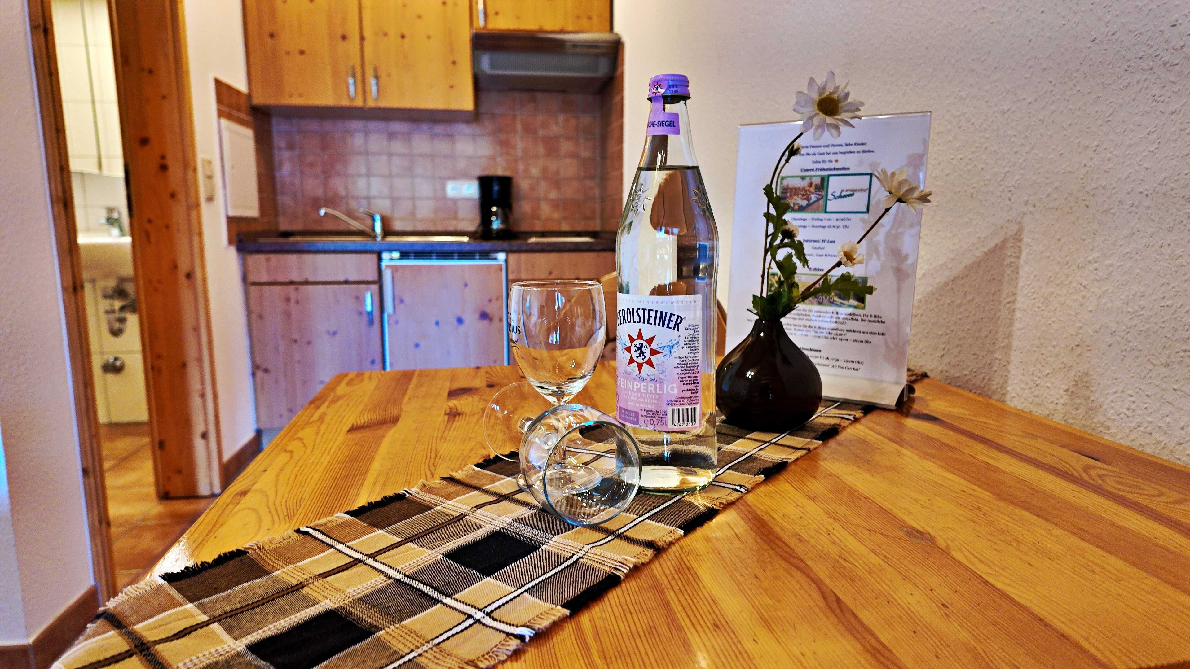 Een houten tafel met een fles drank, een glas en een placemat in een kamer met houten kastjes en een betegelde achterwand.
