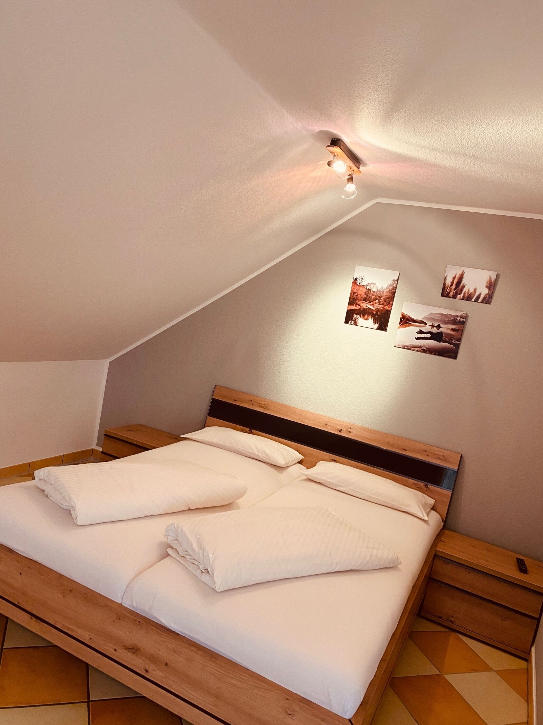 A neatly made bed with white linens in a room with a sloped ceiling and wooden headboard. There are three framed pictures on the wall above the bed.