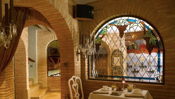 Un comedor con una mesa dispuesta para dos, una araña y un ventanal de vidrio artístico que ofrece vistas de un edificio.