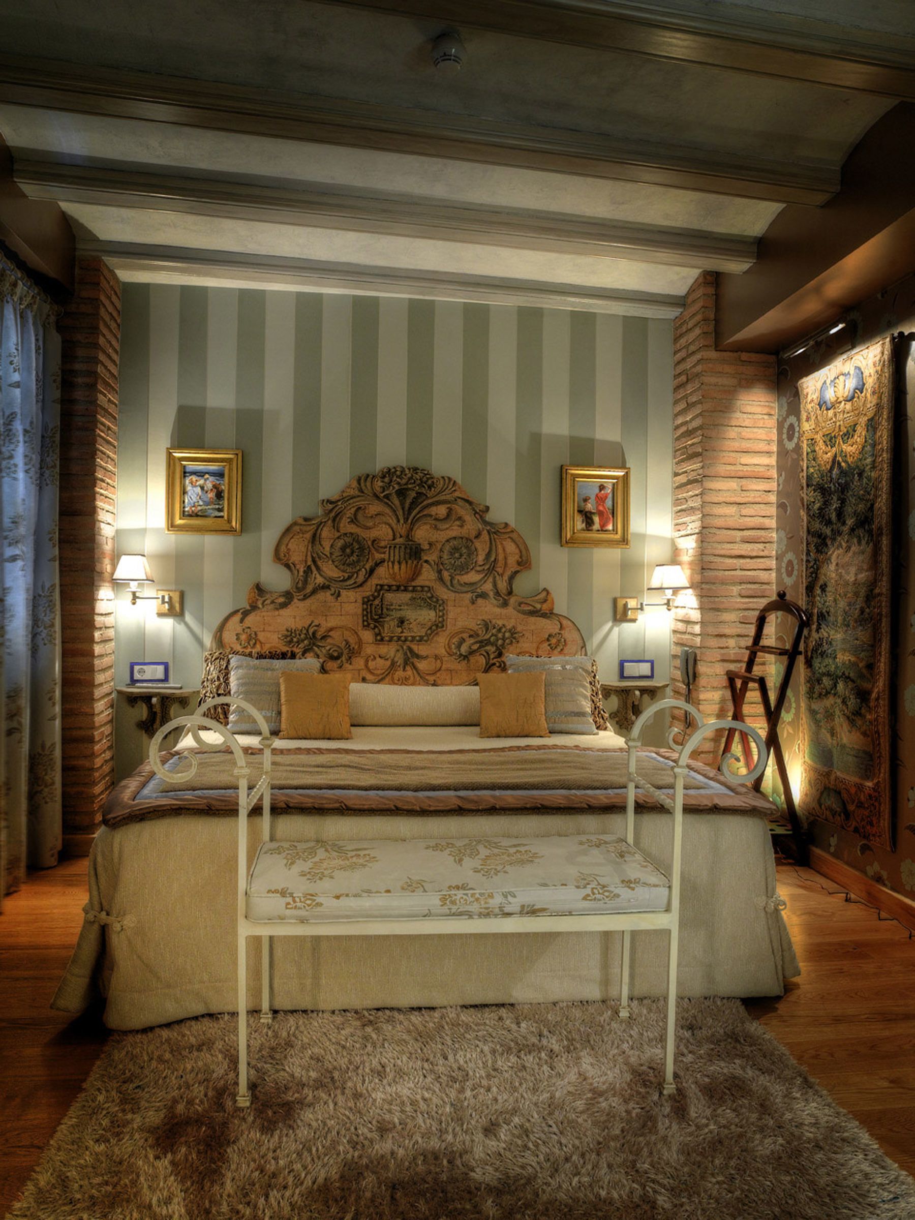Une chambre à coucher avec un grand lit, deux lampes de chevet, une tête de lit ornée de motifs complexes, une petite table et une grande tapisserie au mur.