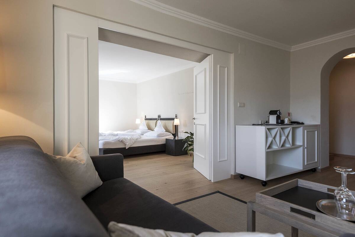 Quarto de hotel moderno com sofá cinzento, cama com roupa de cama branca, pequena cozinha e área de refeições com mesa de vidro.