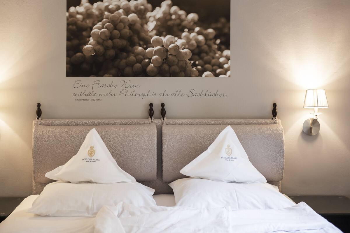 Quarto de hotel com cama, duas almofadas com brasão, luminária na parede e uma moldura com citação sobre a cama.
