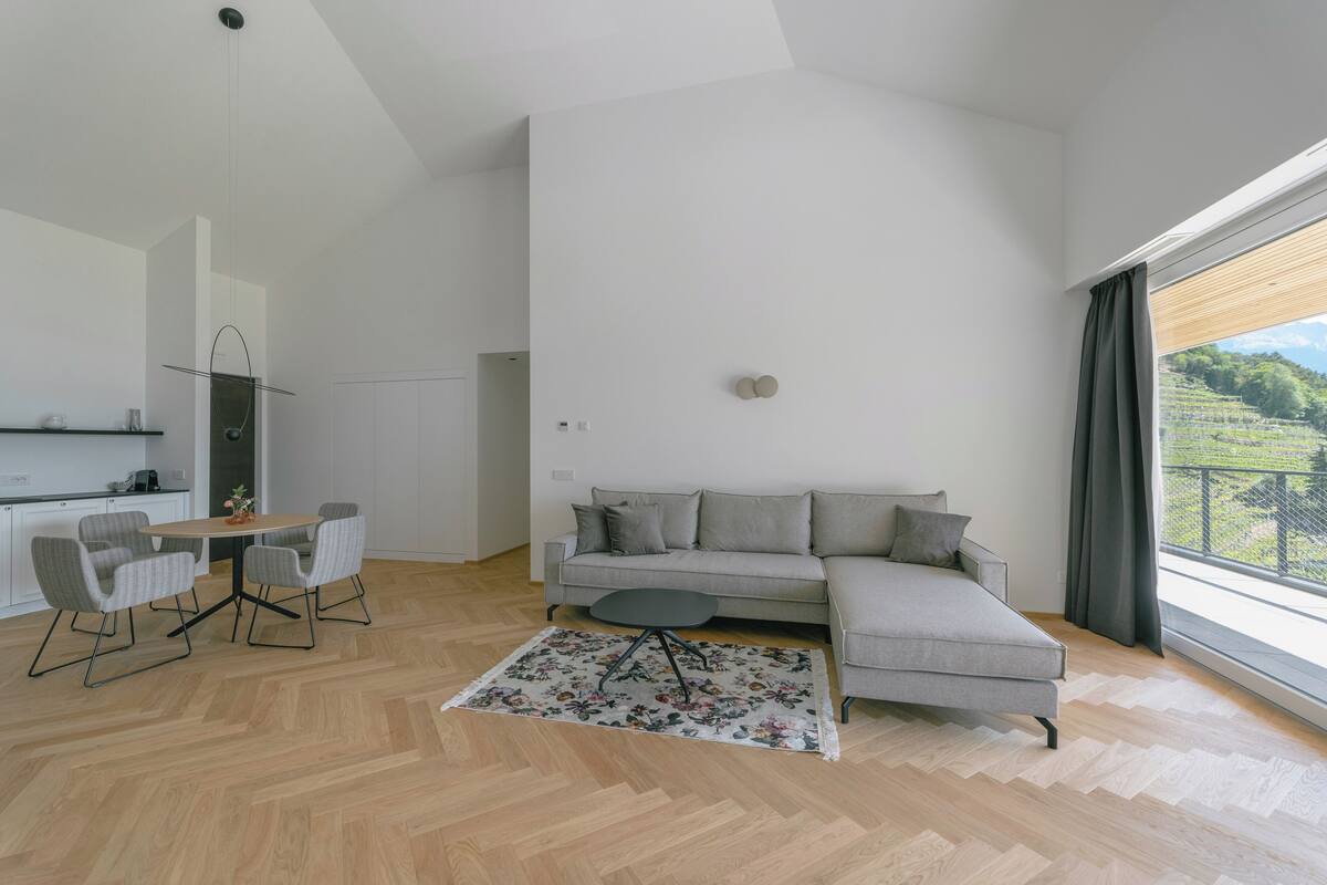 Uma sala de estar moderna com um sofá seccional cinza, uma mesa de jantar redonda com quatro cadeiras e piso de madeira em padrão herringbone.