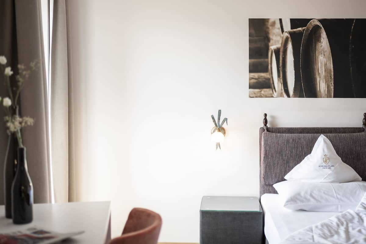 Quarto de hotel com uma cama, uma mesa, uma cadeira, um vaso com flores e um quadro na parede retratando barris.