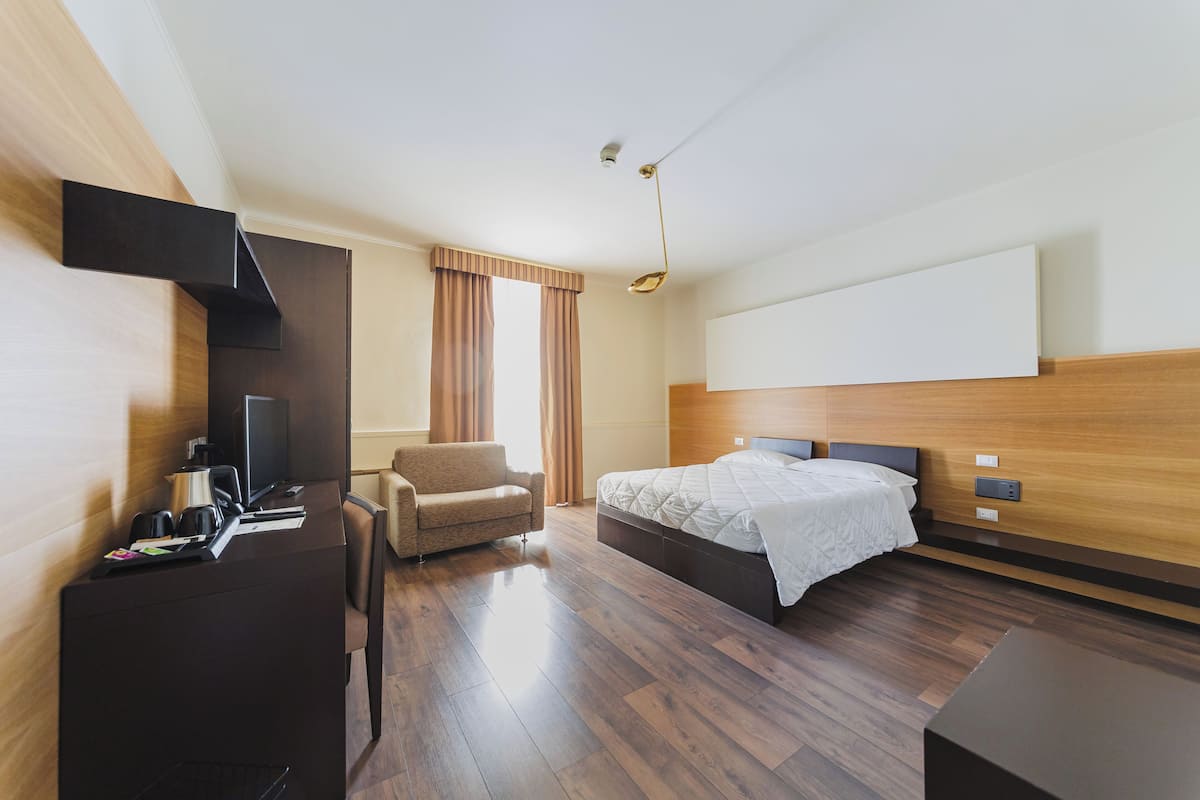Een moderne hotelkamer met een houten vloer, een bed met wit beddengoed, een bureau met een computer en een stoel, en een groot raam met gordijnen.