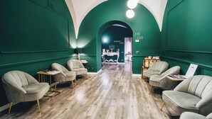 A room with green walls, wooden flooring, and four grey armchairs arranged around a central table. A chandelier hangs from the ceiling, and a sign reads "TEA ROOM". - Hotel San Nicola (Altamura)