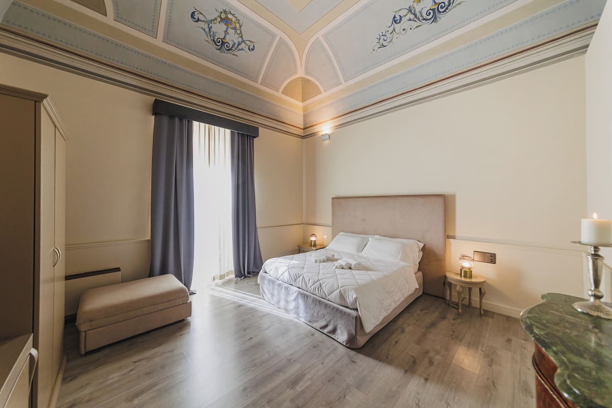 Een slaapkamer met een groot bed, een nachtkastje met een lamp, een bankje en een decoratief plafond.