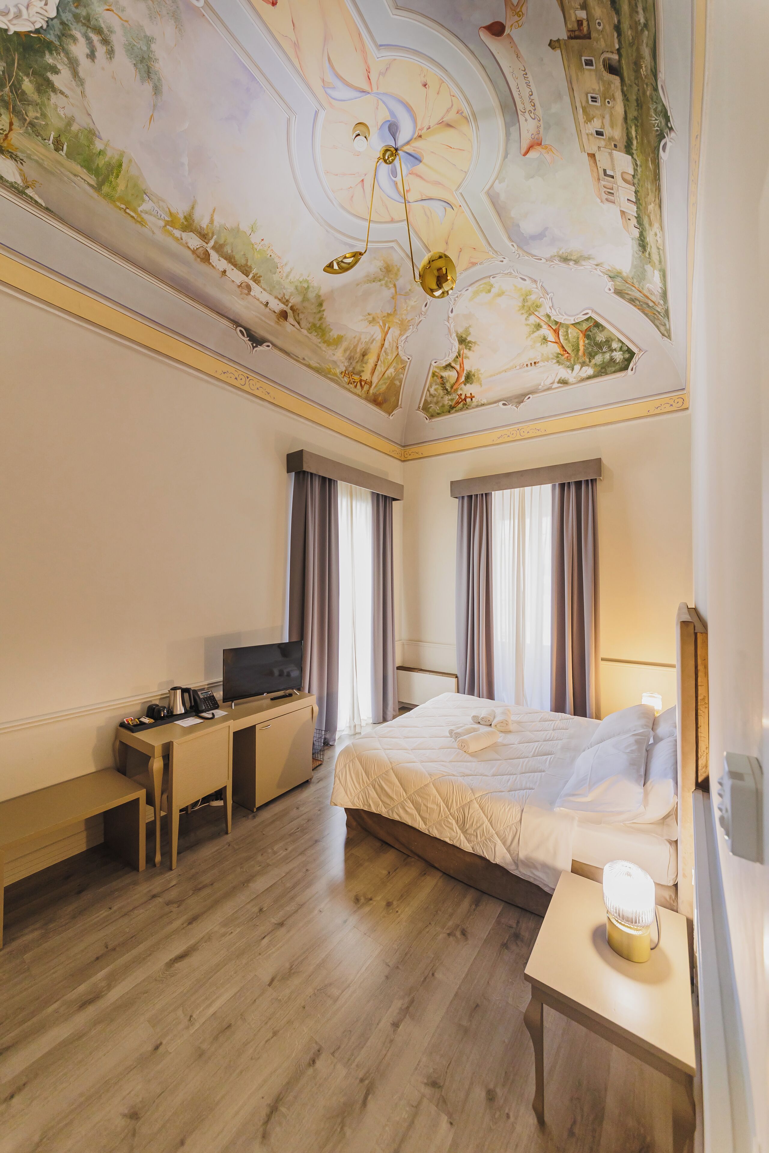 Une chambre d’hôtel comprenant un lit, un bureau avec une télévision, une table de chevet avec une lampe et une fresque au plafond.