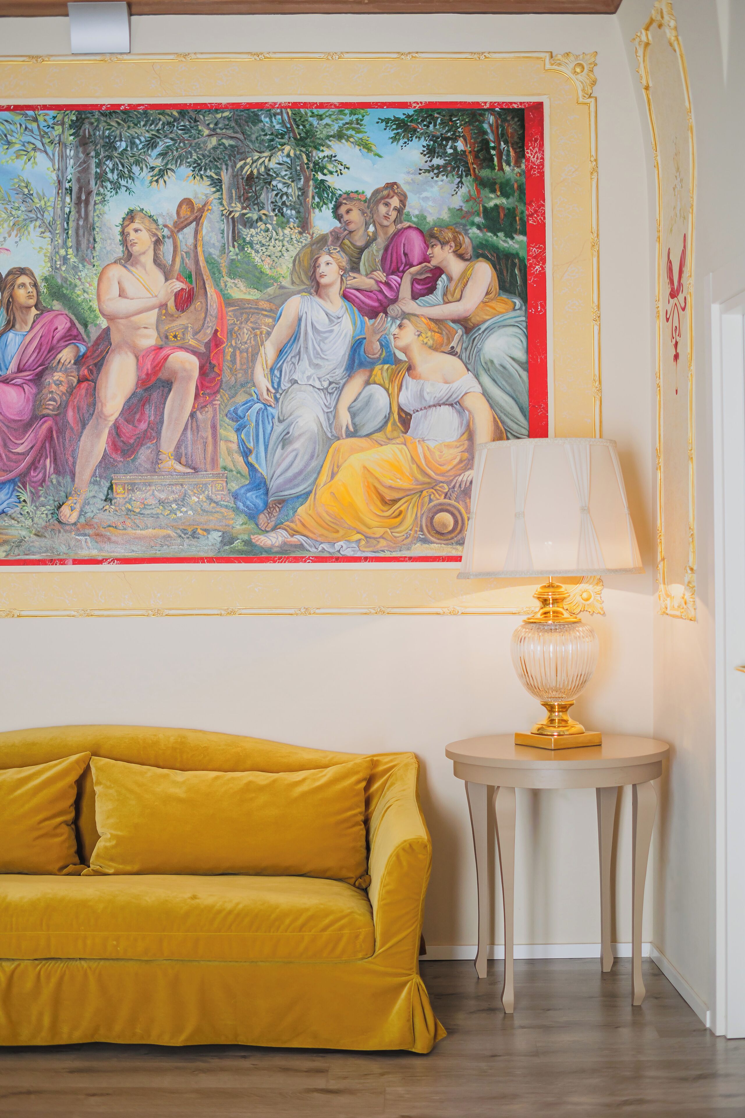 A room with a yellow sofa, a table with a lamp, and a classical painting on the wall.