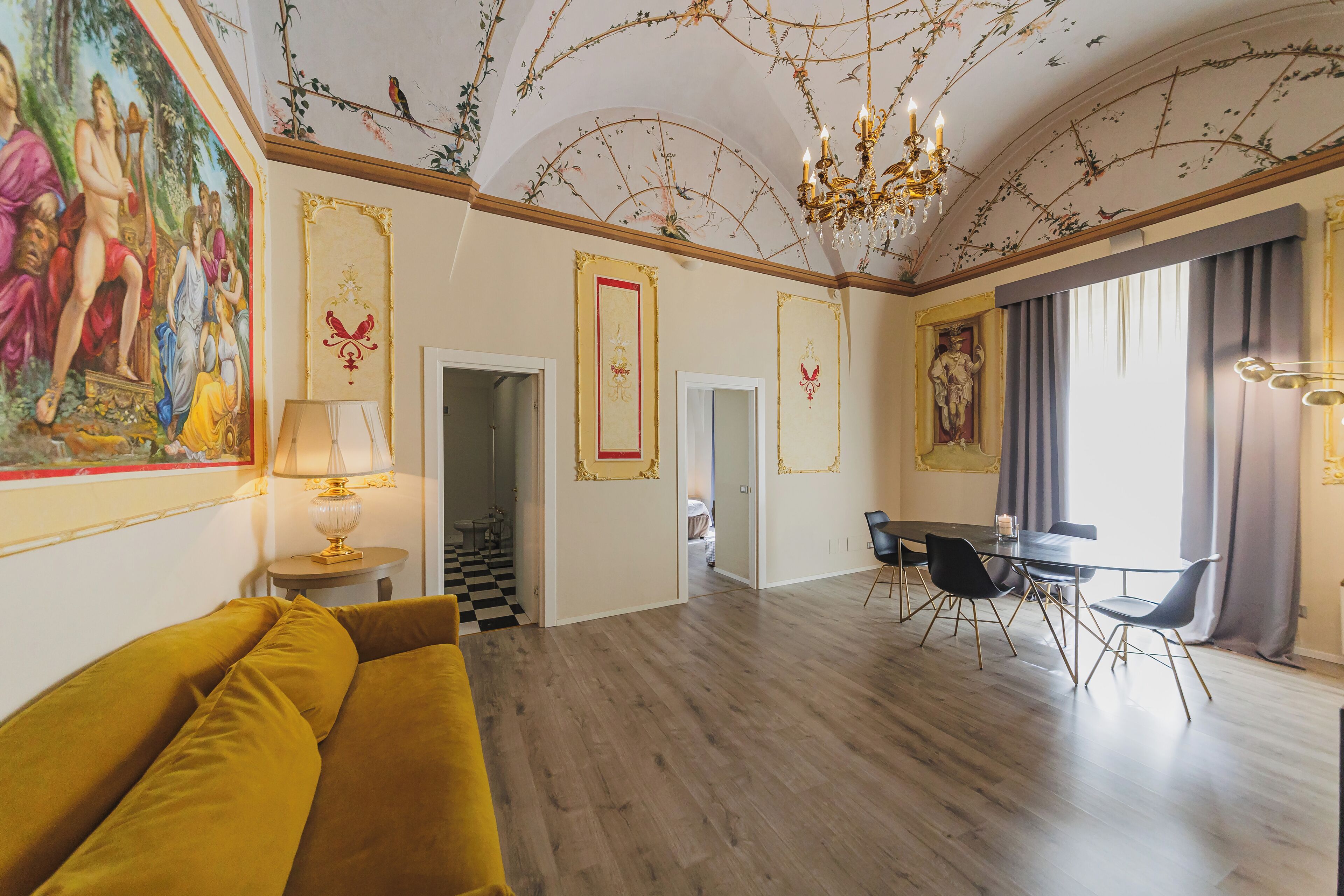 Una habitación amplia con un sofá amarillo, una mesa de comedor con sillas, una araña y un mural grande en la pared.
