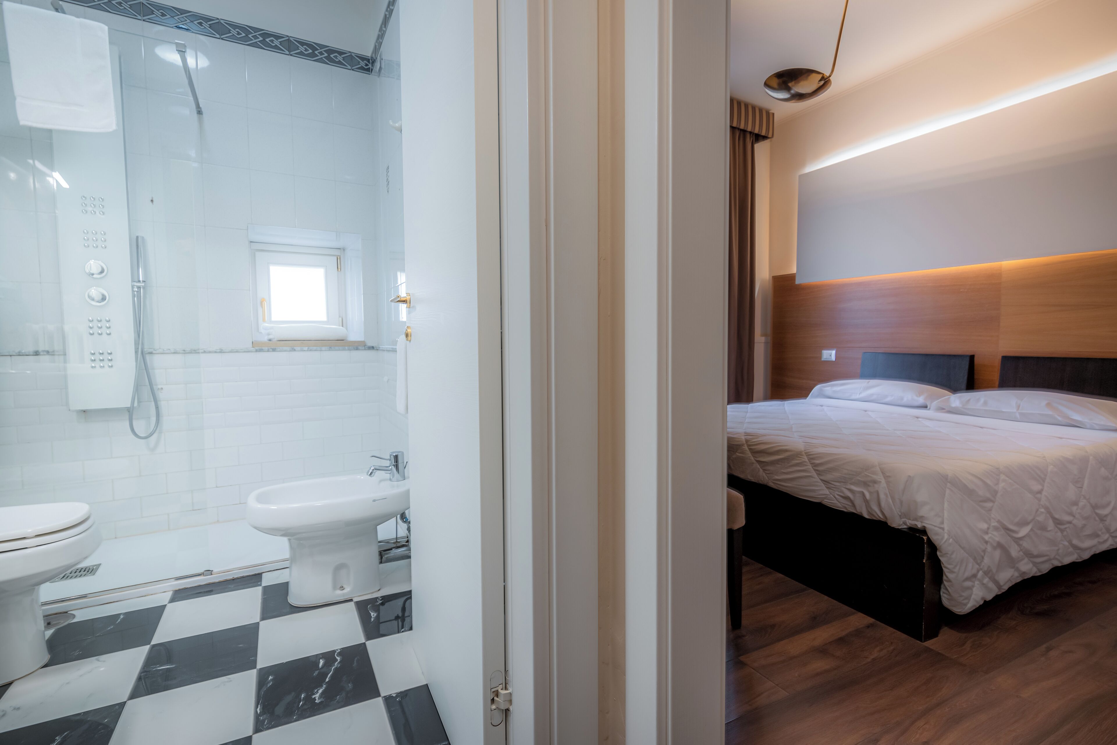 Ein Hotelzimmer mit Schachbrettmuster-Boden, einem Bett, einem Nachttisch und einem Badezimmer mit Toilette, Bidet und Dusche.