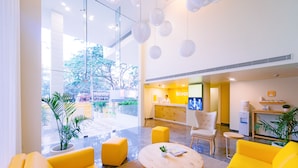 A bright reception area with yellow furniture, a reception desk, and a large window offering a view of the outdoors. - Bloom Hotel - Indiranagar  (Bengaluru)