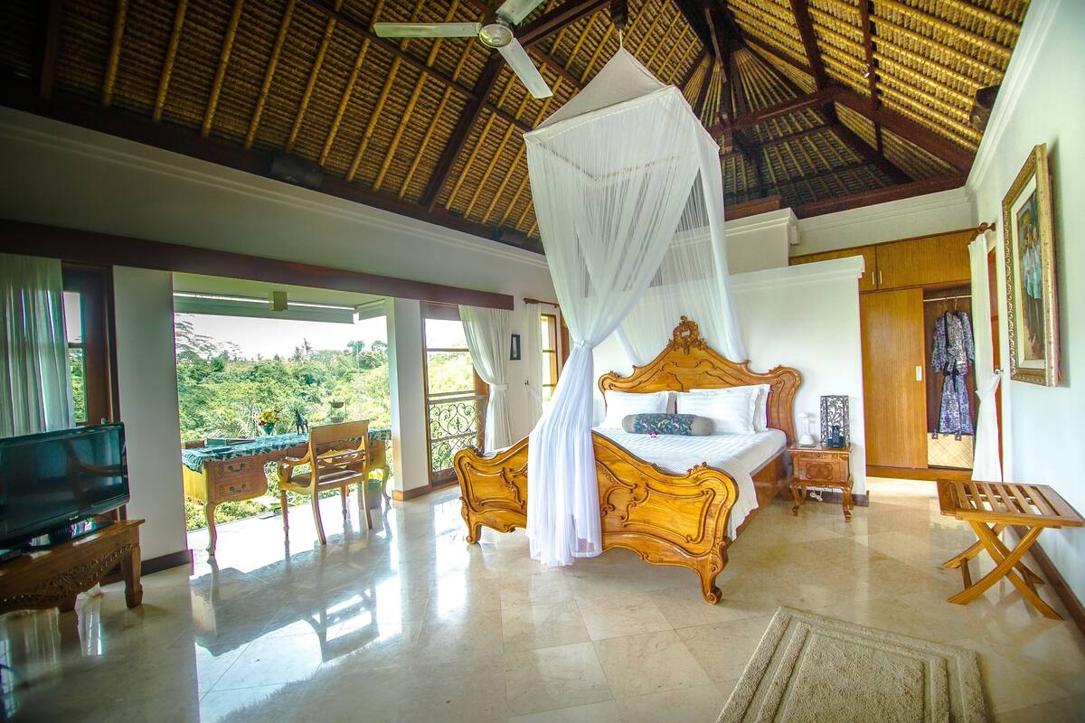 A spacious bedroom with a large bed, a television, and a balcony with a view of greenery.