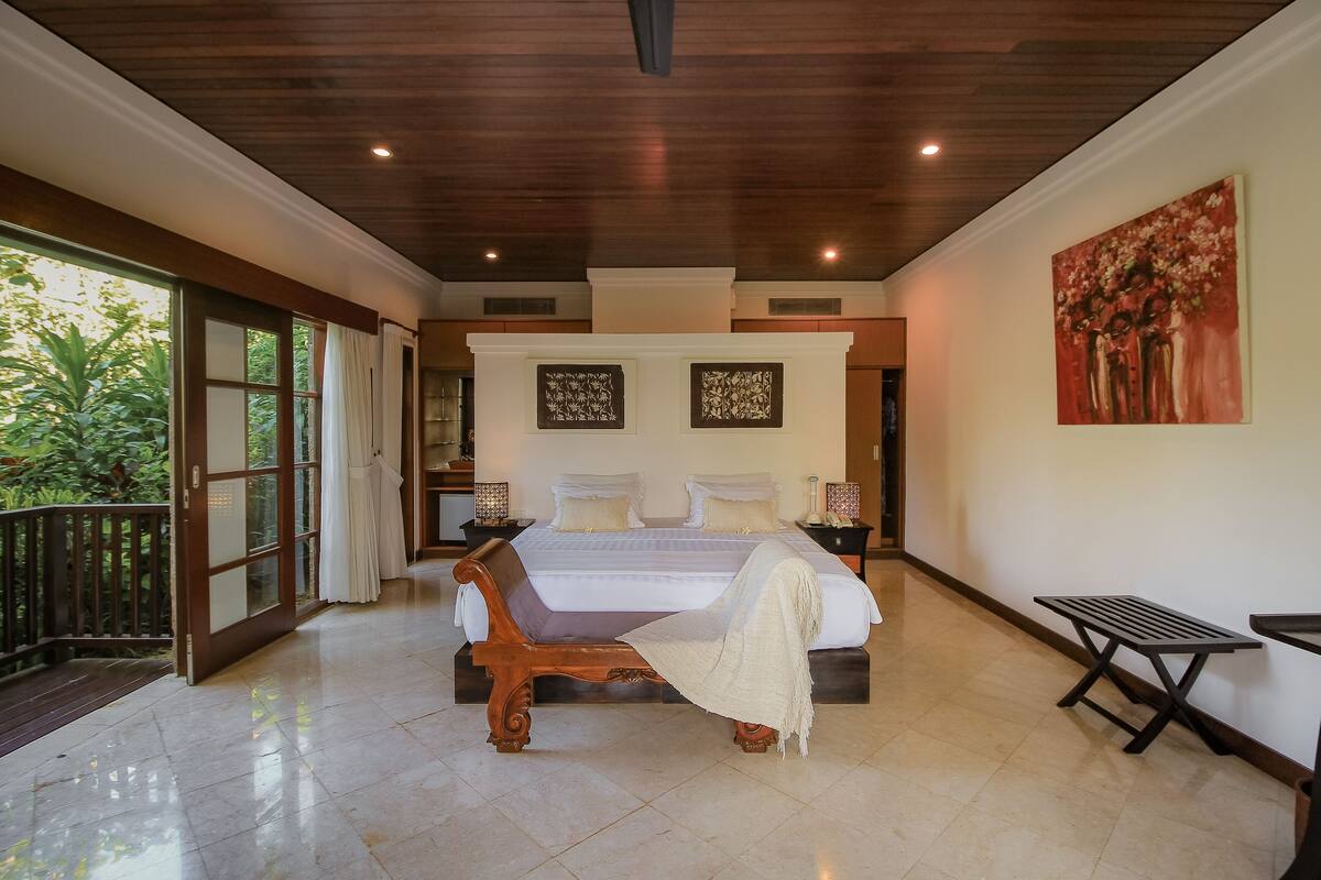 A spacious bedroom with a large bed, a bench, and a view of greenery outside.