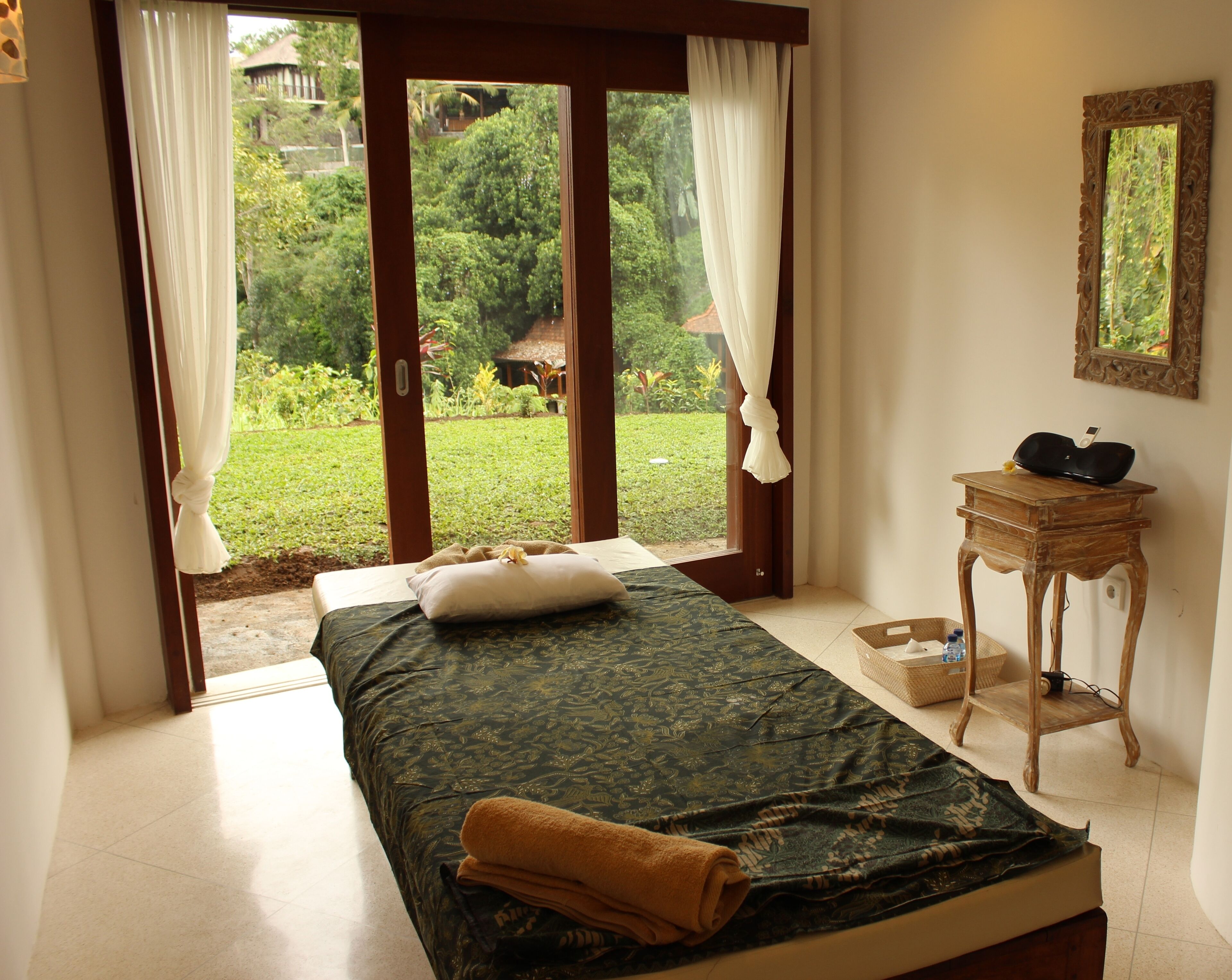 Quarto com cama, mesa de cabeceira, espelho e vista para um jardim através de uma porta de vidro.