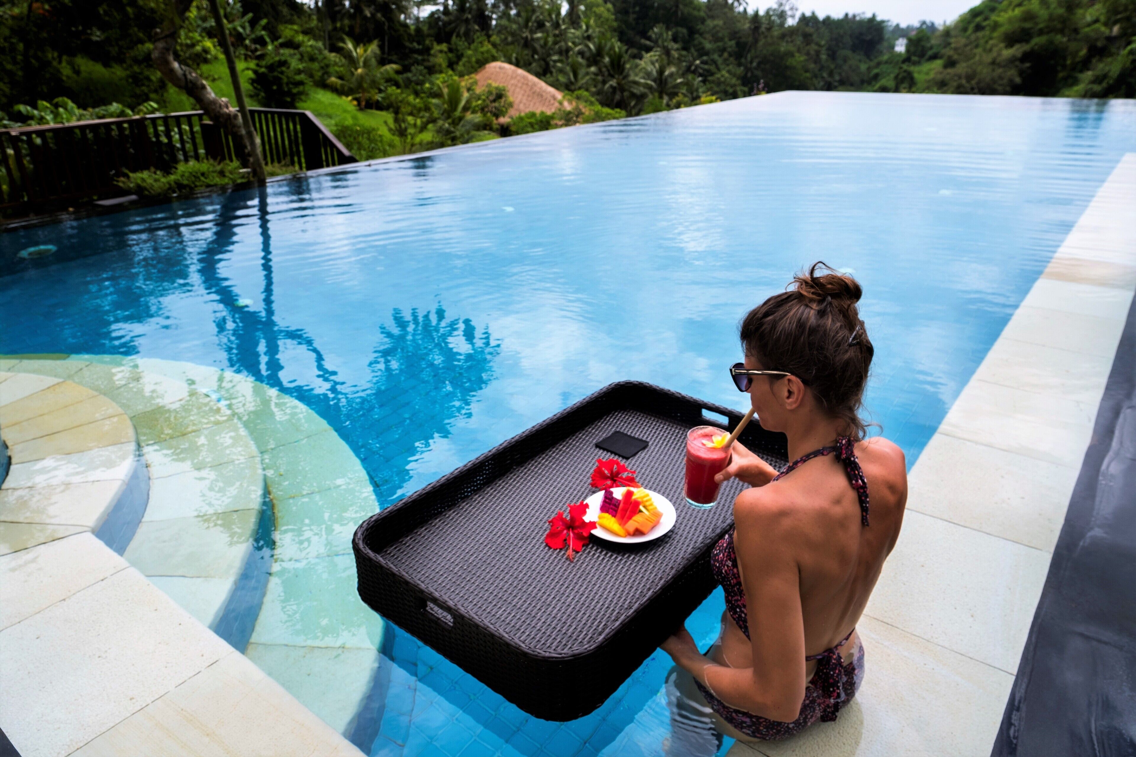 Una persona sosteniendo una bandeja con una bebida y fruta junto a una piscina con agua azul cristalina.