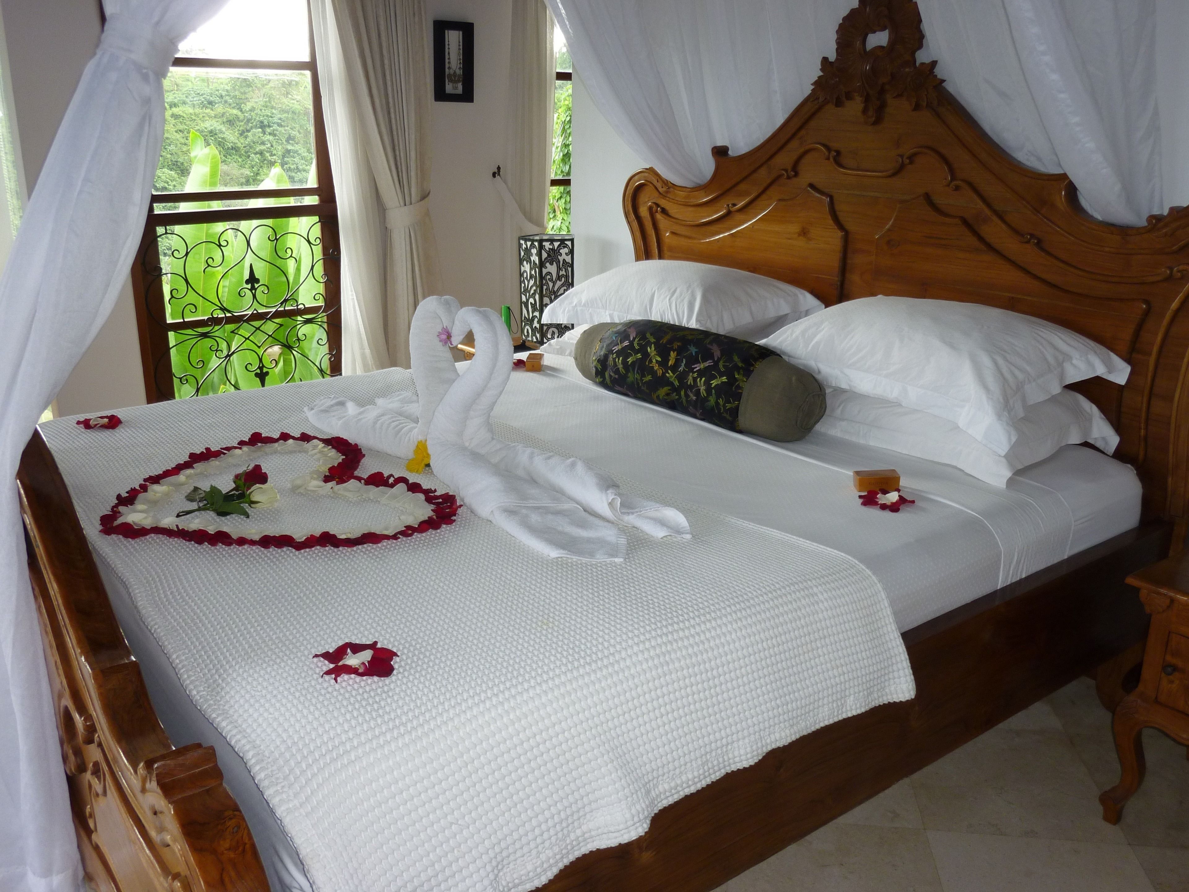 A neatly made bed with a white bedspread, a heart-shaped arrangement of rose petals, and a decorative pillow.