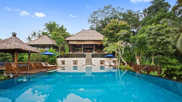 A clear view of a swimming pool with lounge chairs and umbrellas, surrounded by lush greenery and a thatched-roof building. - Amori Villas (Ubud)