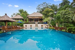 A clear view of a swimming pool with lounge chairs and umbrellas, surrounded by lush greenery and a thatched-roof building. - Amori Villas (Ubud)