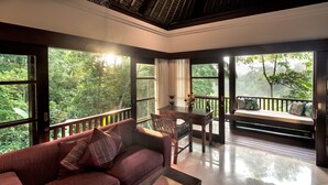 A room with a wooden floor, a sofa with cushions, a table with chairs, and large windows offering a view of greenery. - Amori Villas (Ubud)