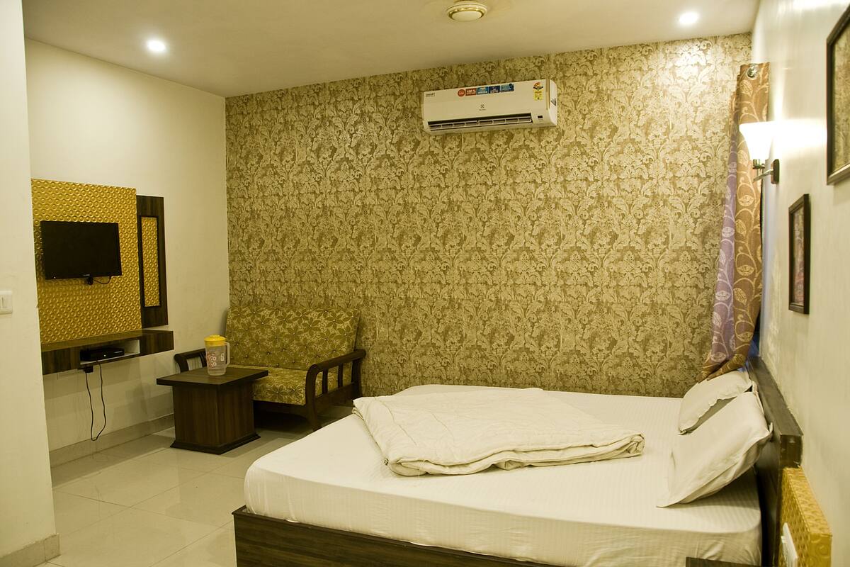 A hotel room with a bed, a chair, a small table, a TV, and an air conditioning unit.