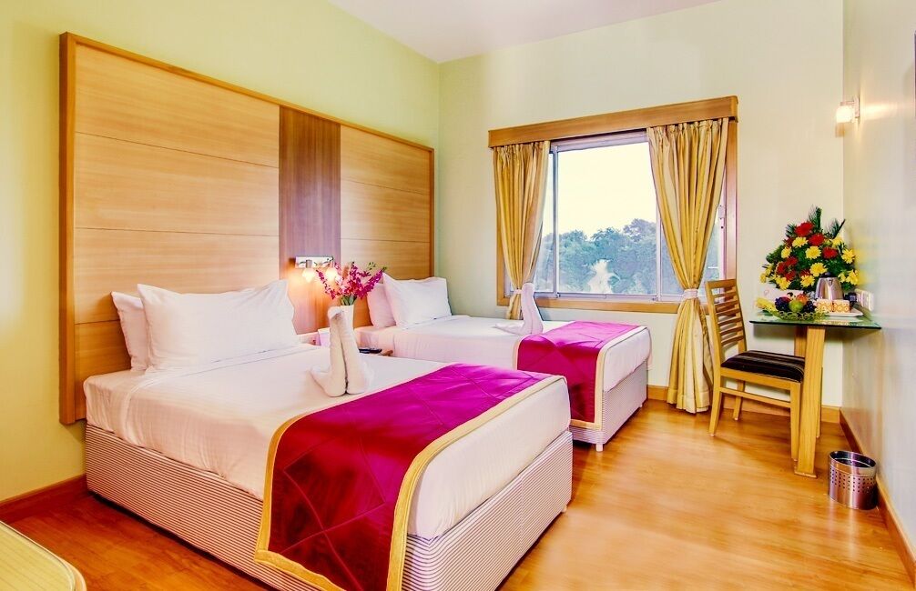 A hotel room with two beds, a wooden headboard, a window with curtains, a small table with a vase of flowers, and a chair.