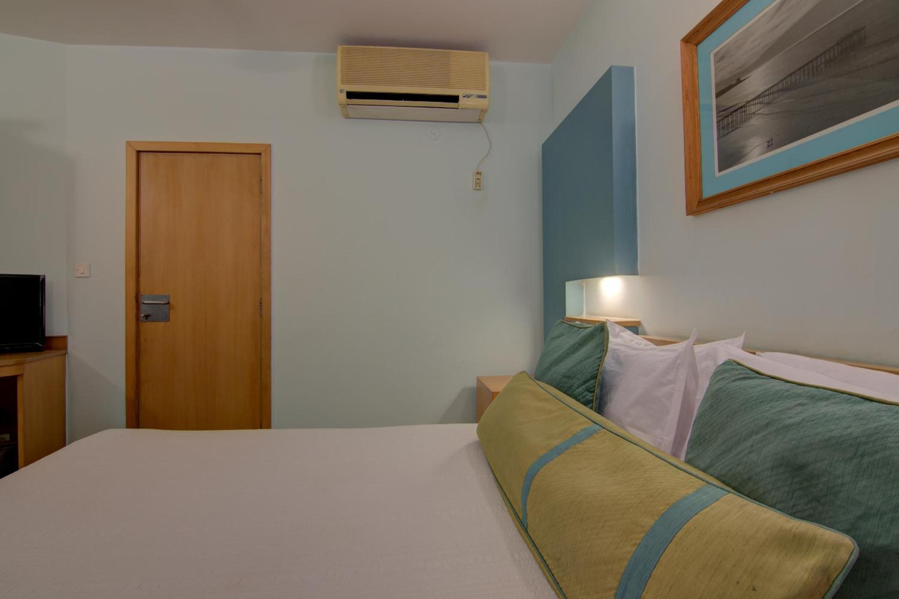 A hotel room with a bed, a nightstand, a television, an air conditioning unit, and a framed picture on the wall.
