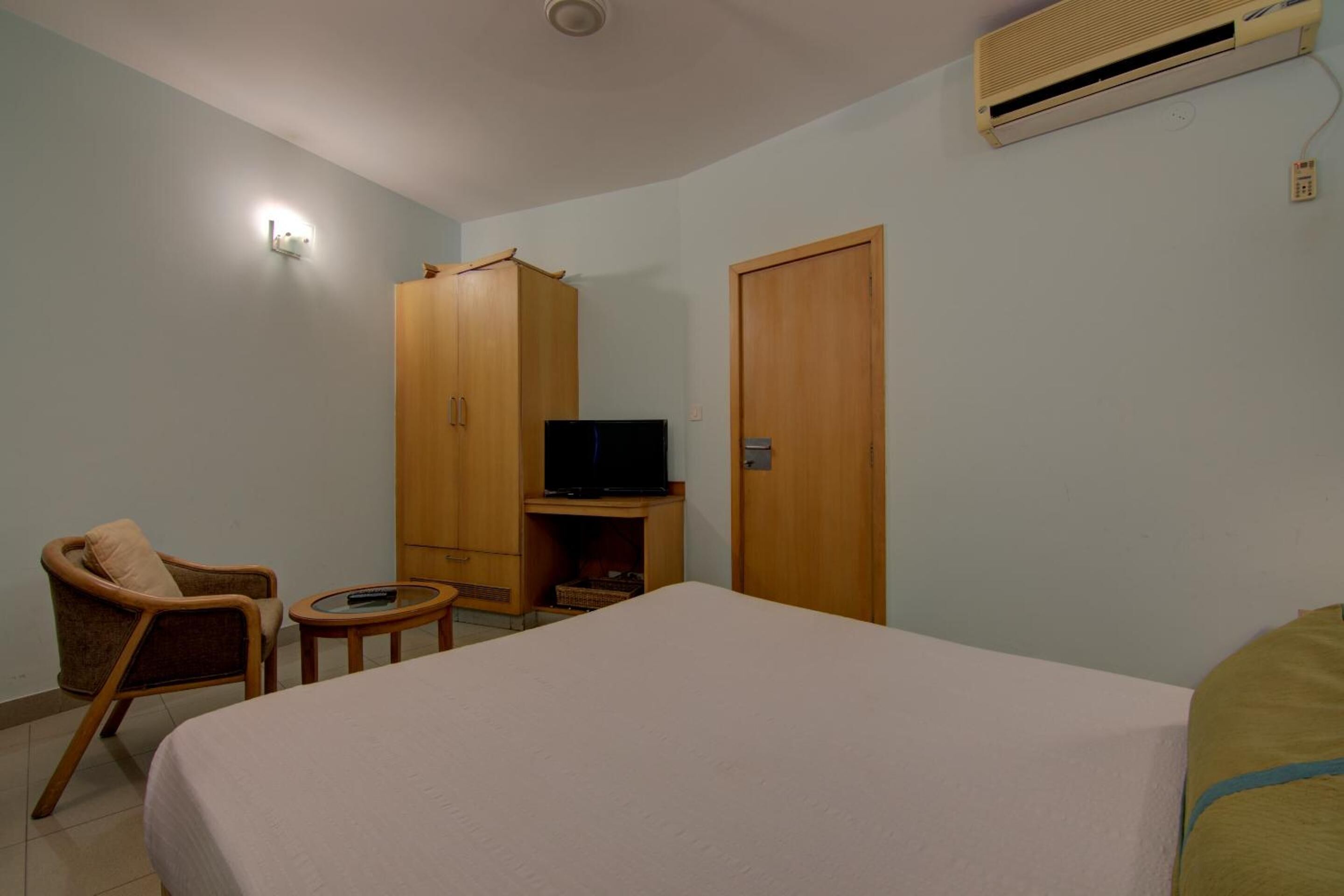 A hotel room with a bed, a television on a stand, a wooden wardrobe, a chair, and a small table.