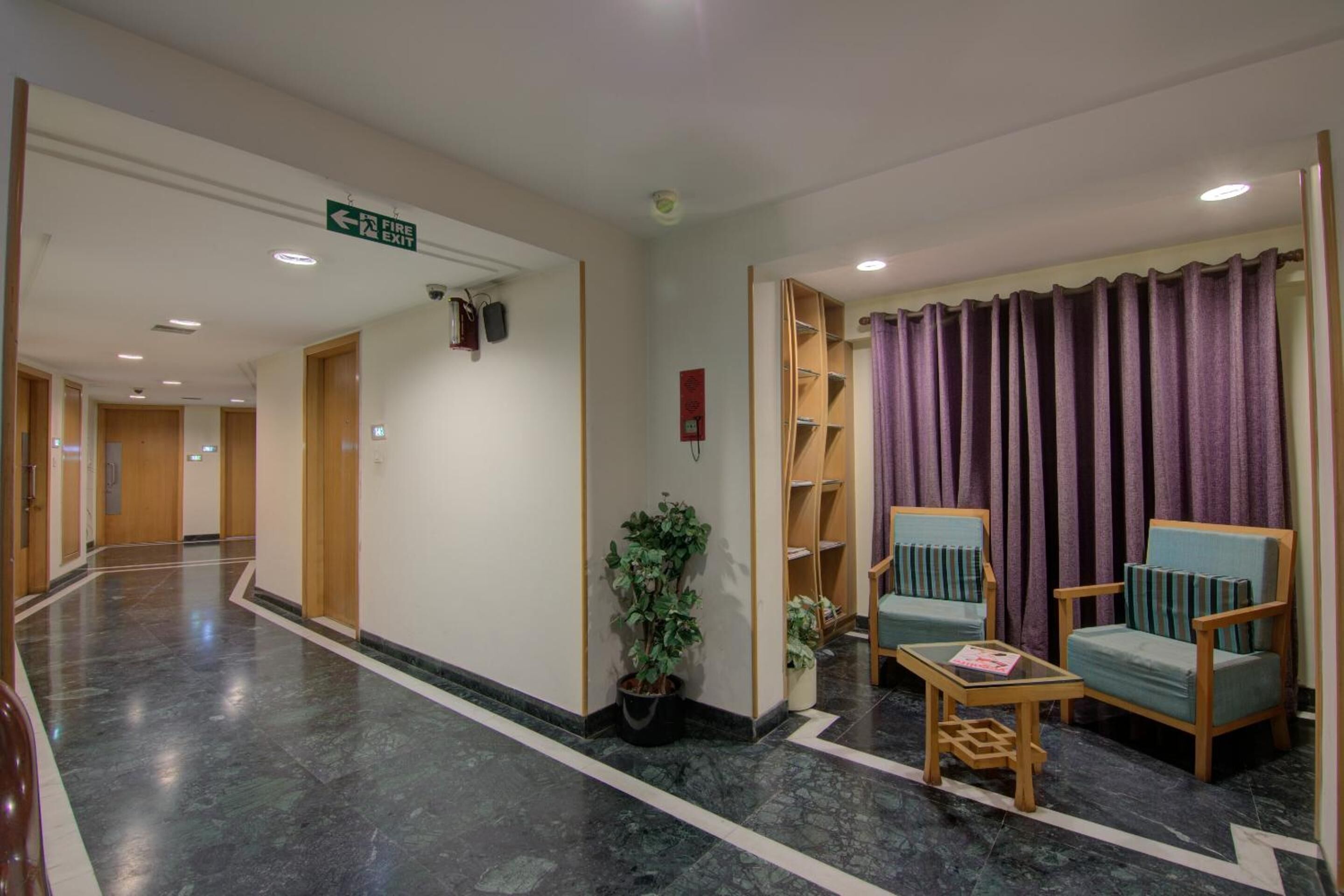 Un'hall dell'hotel con pavimento in marmo, un cartello di uscita di sicurezza, una pianta in vaso e due poltrone con un tavolino.
