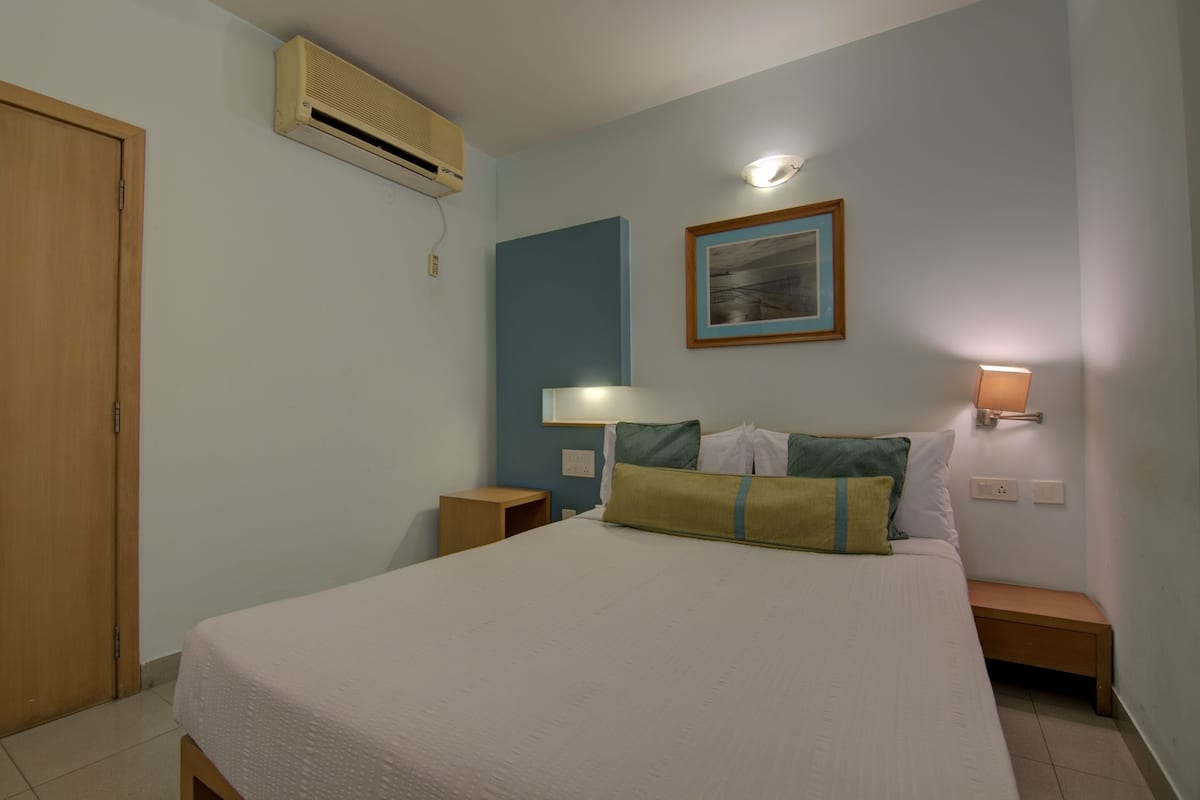 A bedroom with a bed, bedside tables, a wall-mounted air conditioner, a framed picture, and a wall-mounted reading light.
