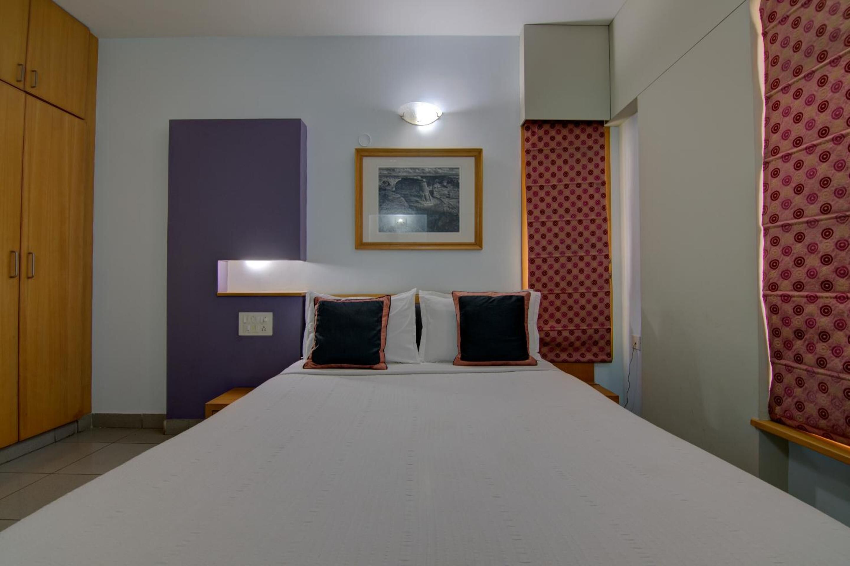 A bedroom with a bed, a wardrobe, a framed picture on the wall, and patterned curtains.