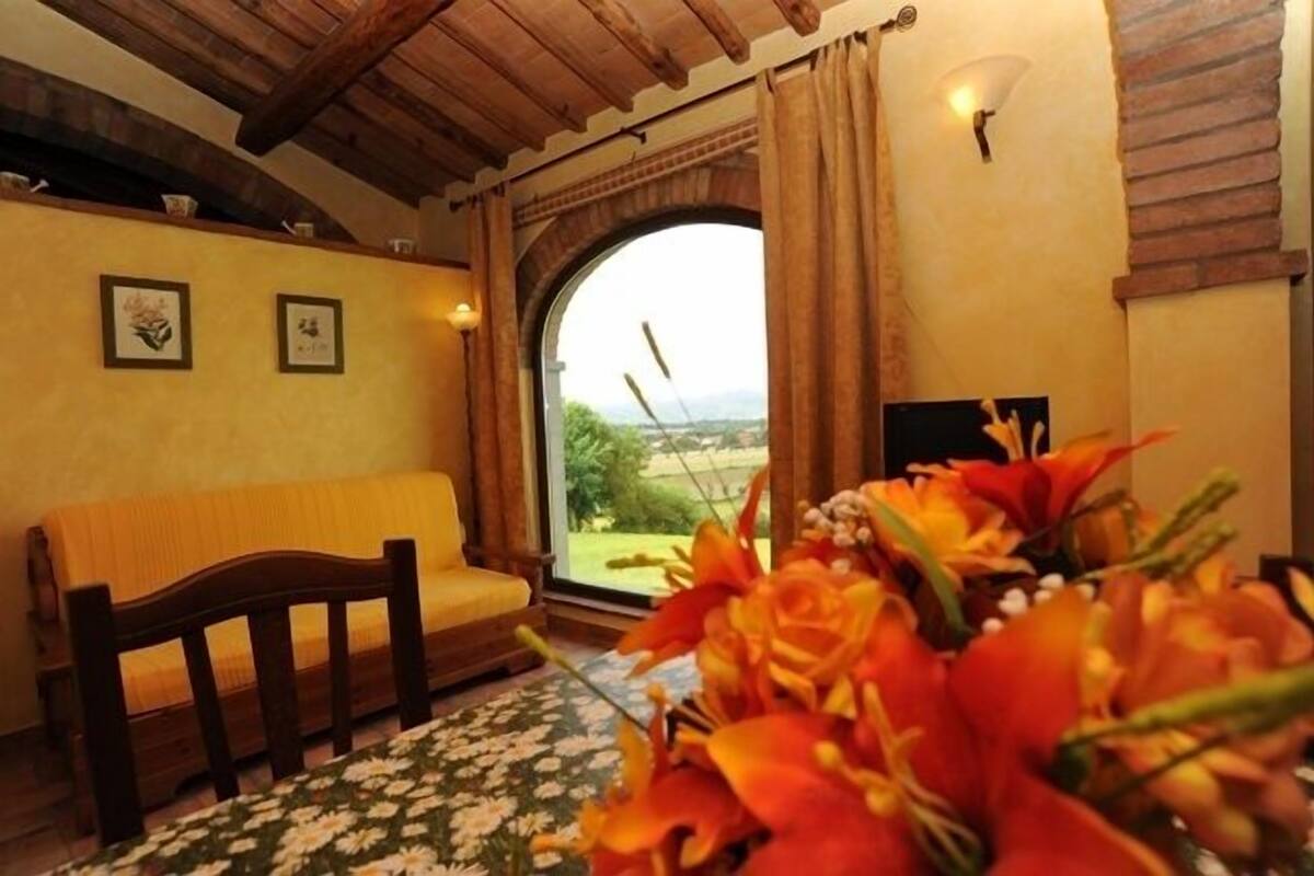 Una habitación con un banco de madera, un ventanal con cortinas y un arreglo floral.