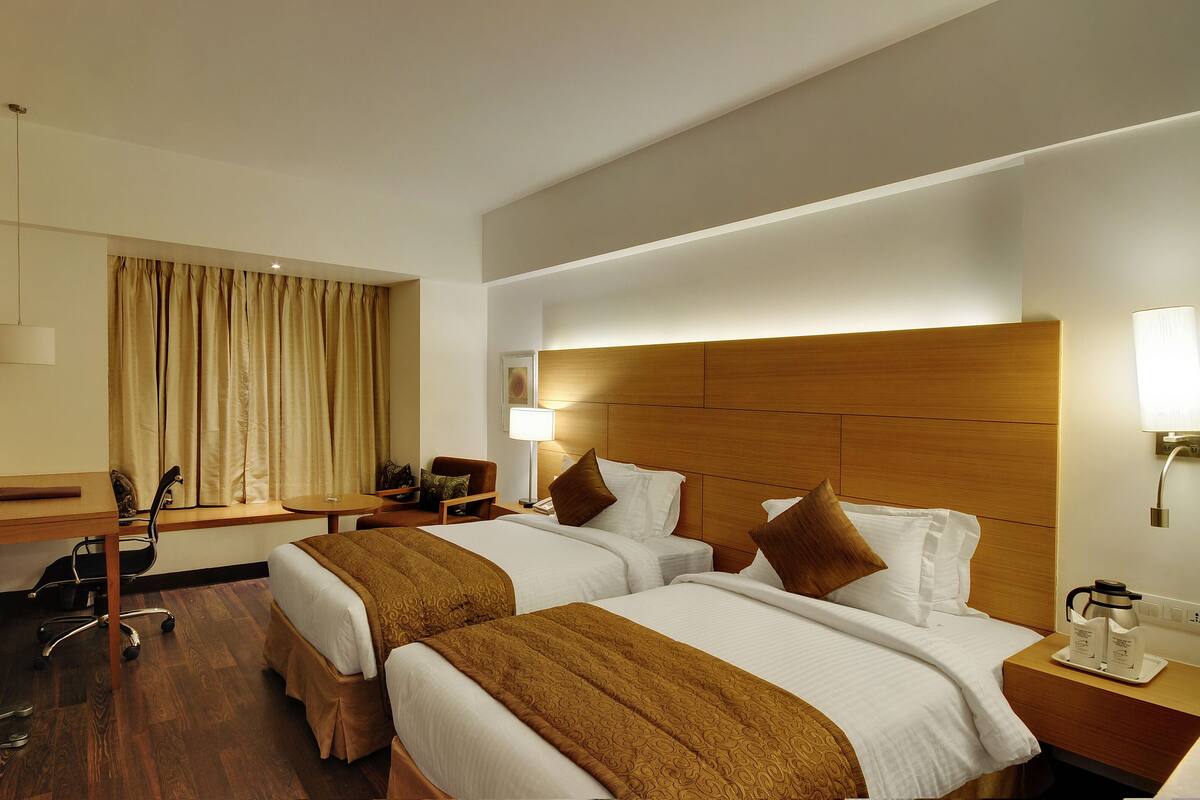 A hotel room with two beds, a desk, a chair, a TV, and a window with curtains.