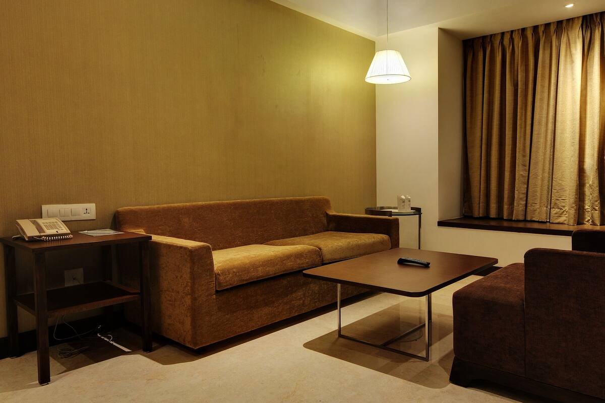 A hotel room with a sofa, a small table, a chair, a lamp, and a window with curtains.