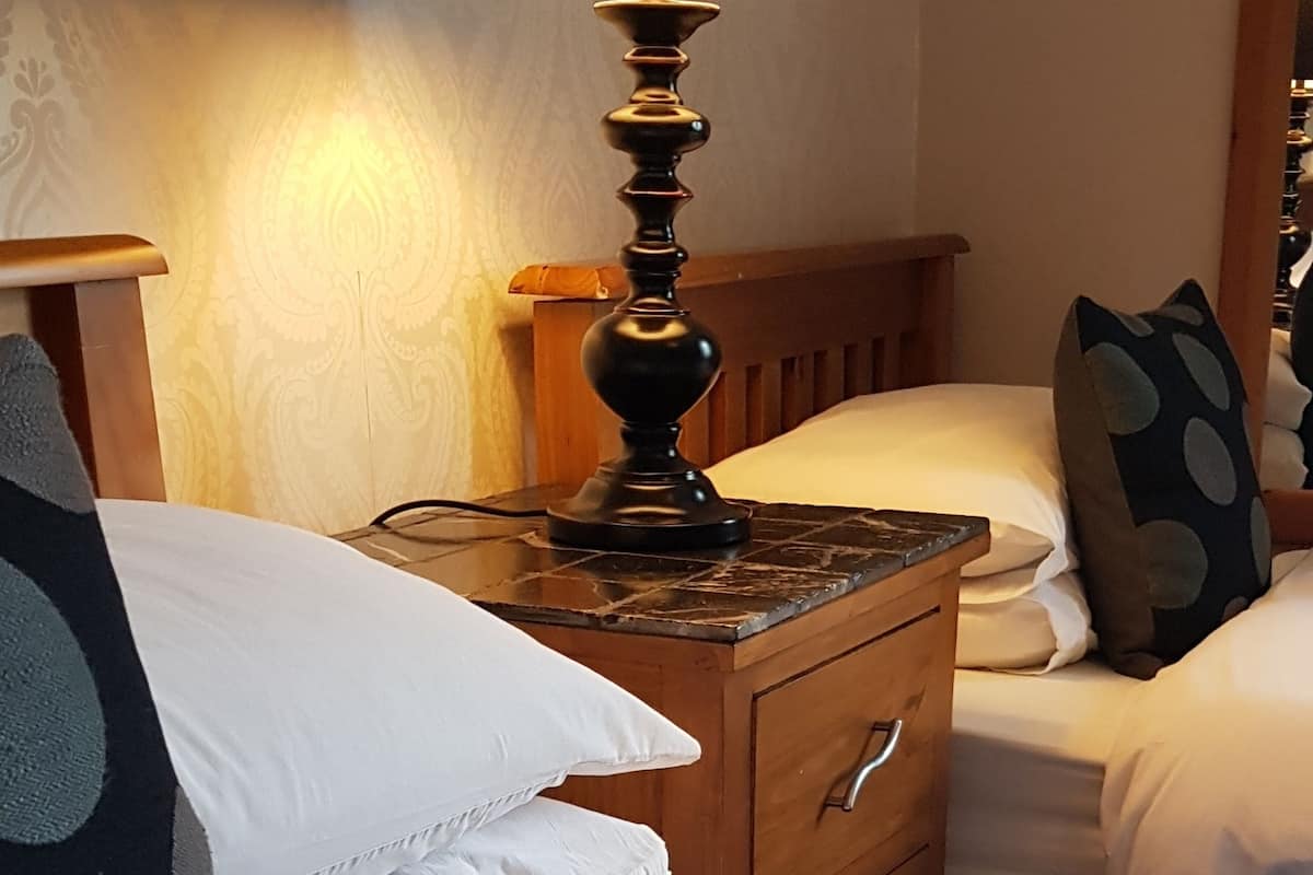 Uma cama bem arrumada com lençóis brancos e detalhes em renda, uma mesa de cabeceira de madeira com abajur, e uma parede decorada ao fundo.