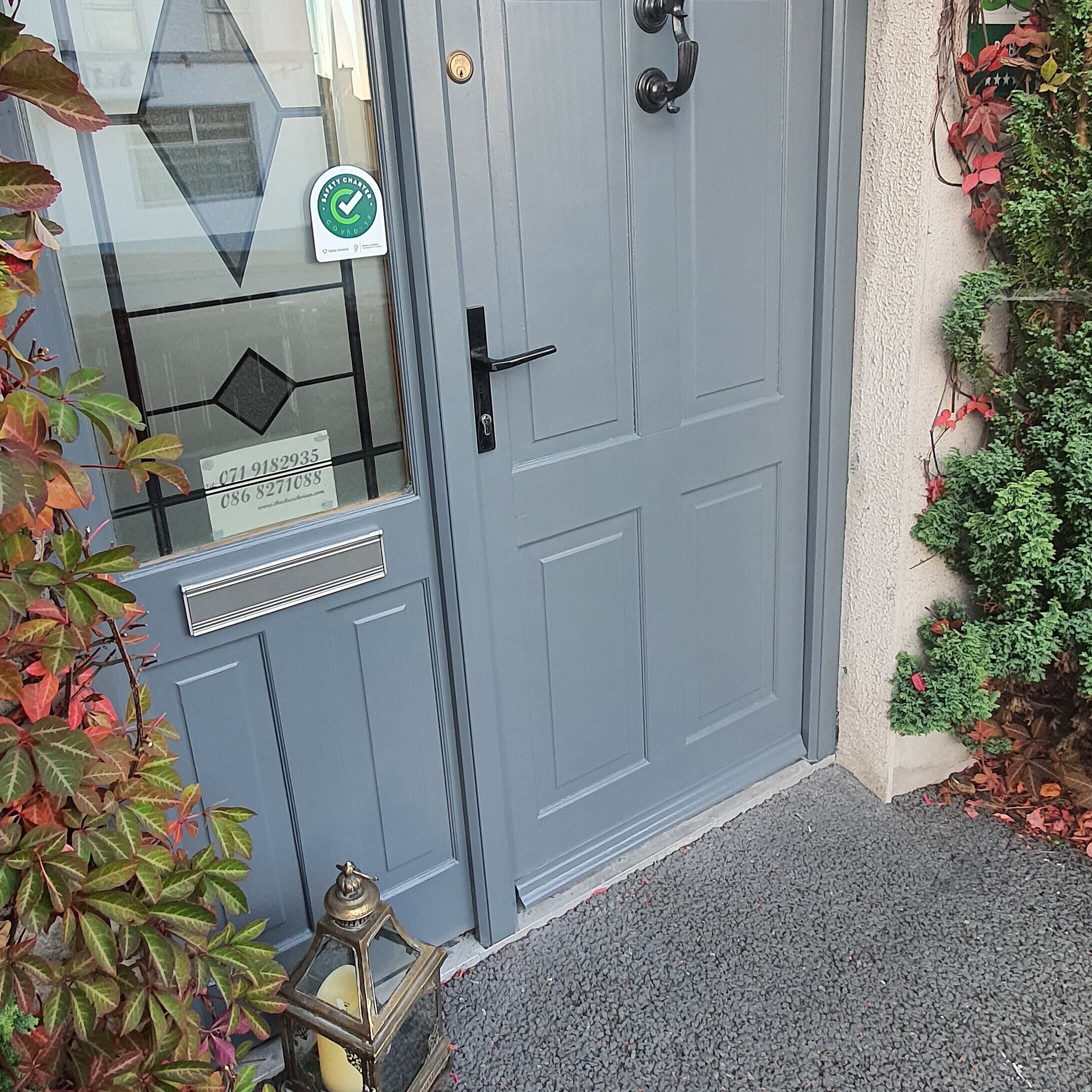 Uma porta cinzenta com corrente de segurança, uma janela com padrão geométrico e uma pequena planta de folhas vermelhas.