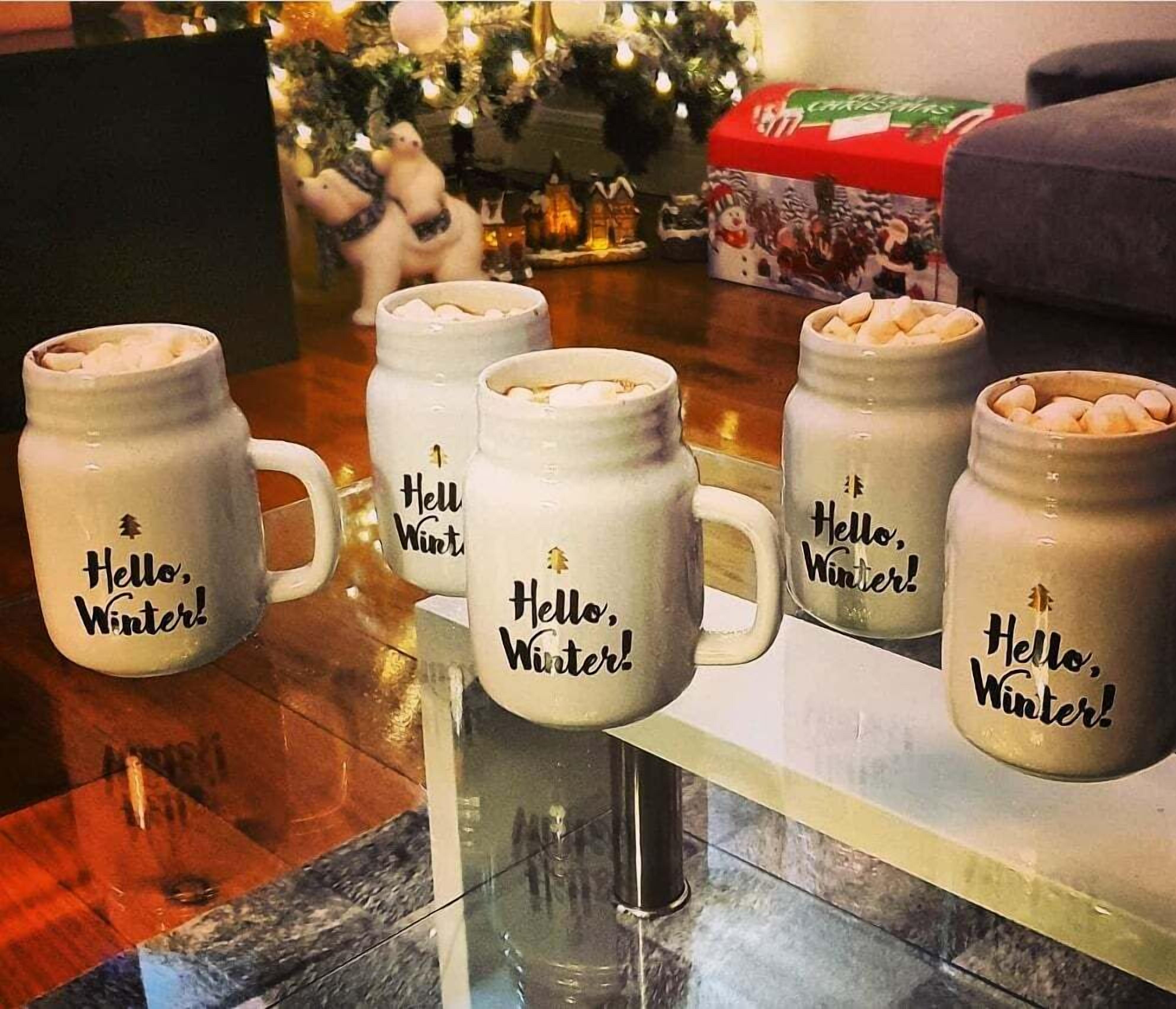 Una mesa con varias tazas blancas que dicen '¡Hola, Invierno!' y un árbol de Navidad al fondo.