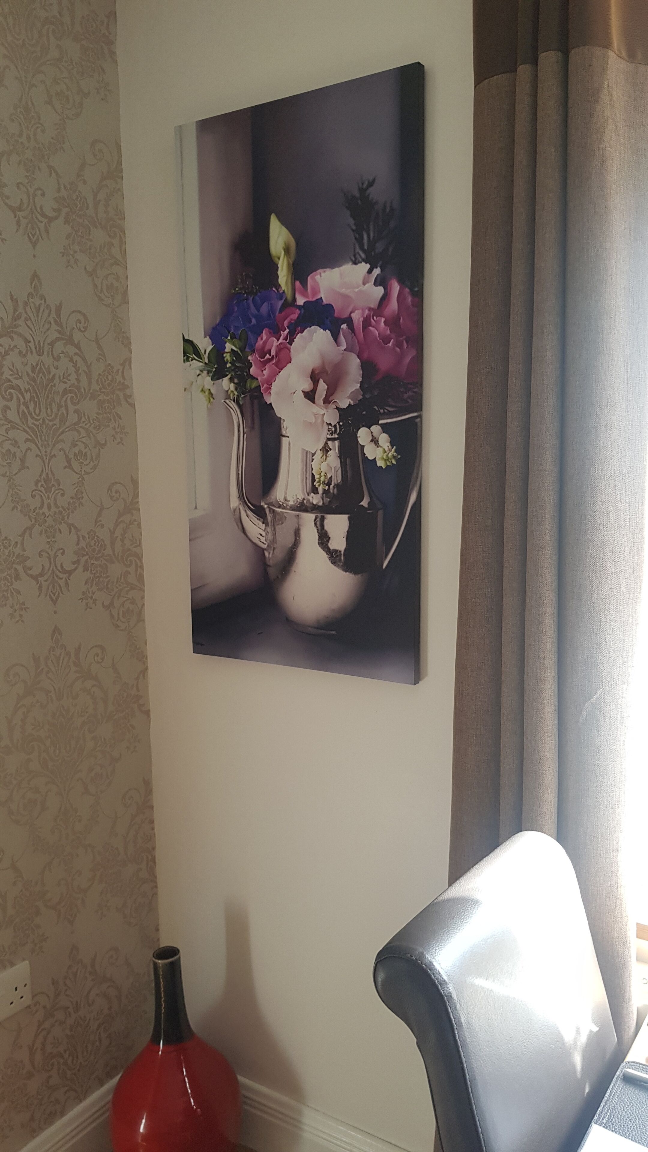 Fotografía en la pared de un jarrón con flores, un jarrón rojo y una silla gris.