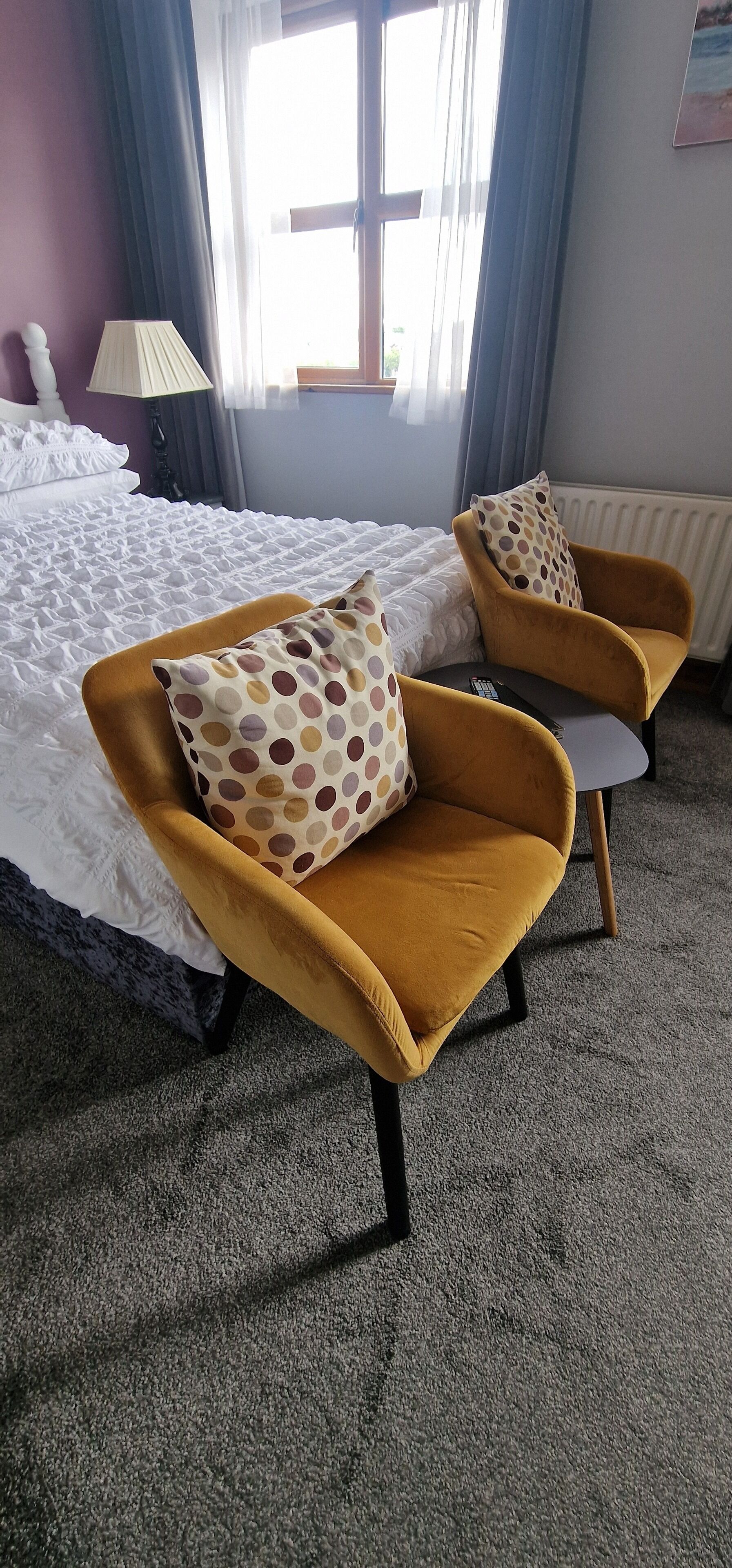 A hotel room with a bed, two armchairs with polka dot cushions, a lamp, and a window with curtains.