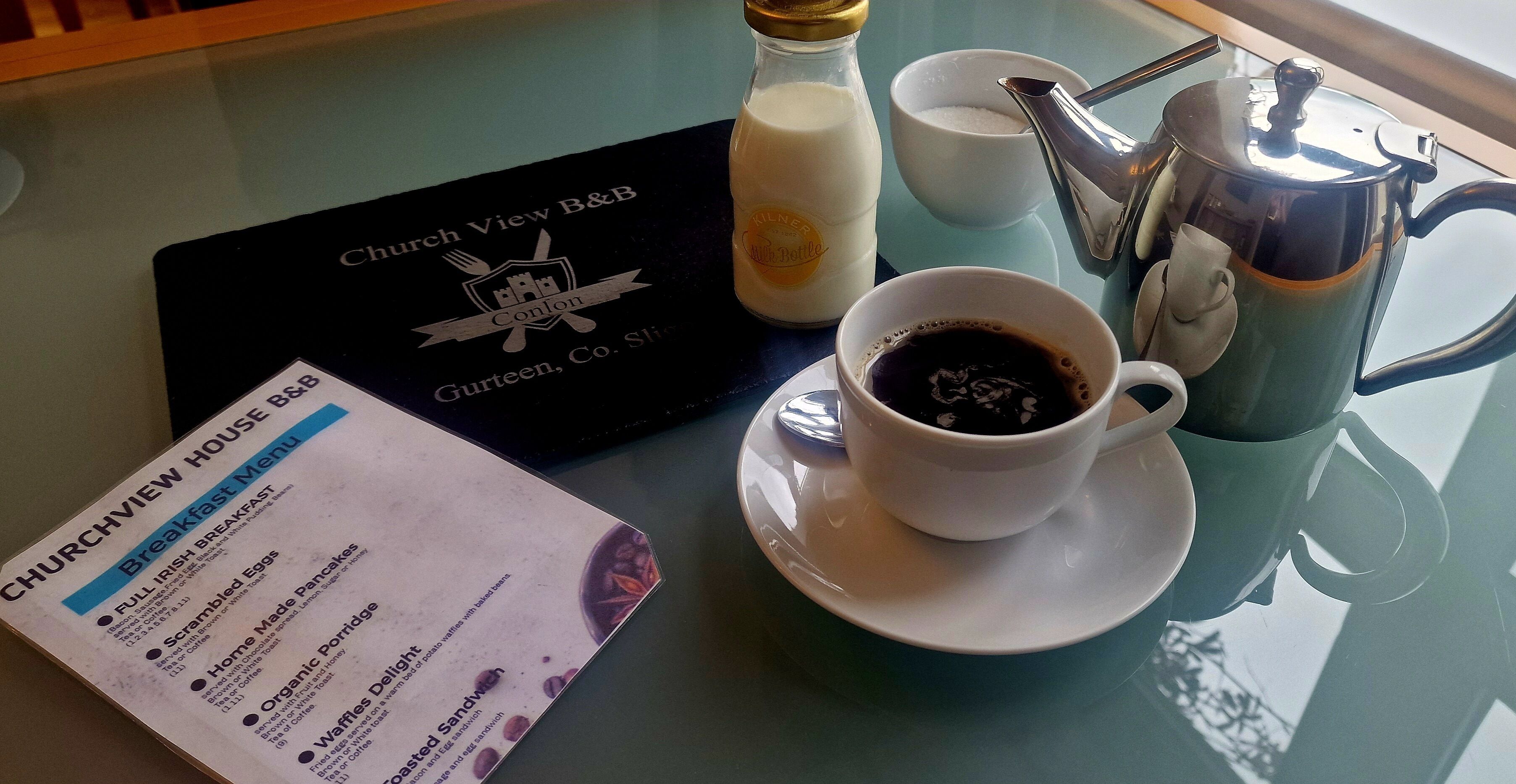Uma mesa de café da manhã com um cardápio, uma xícara de café, um garrafão de leite e um bule de prata.