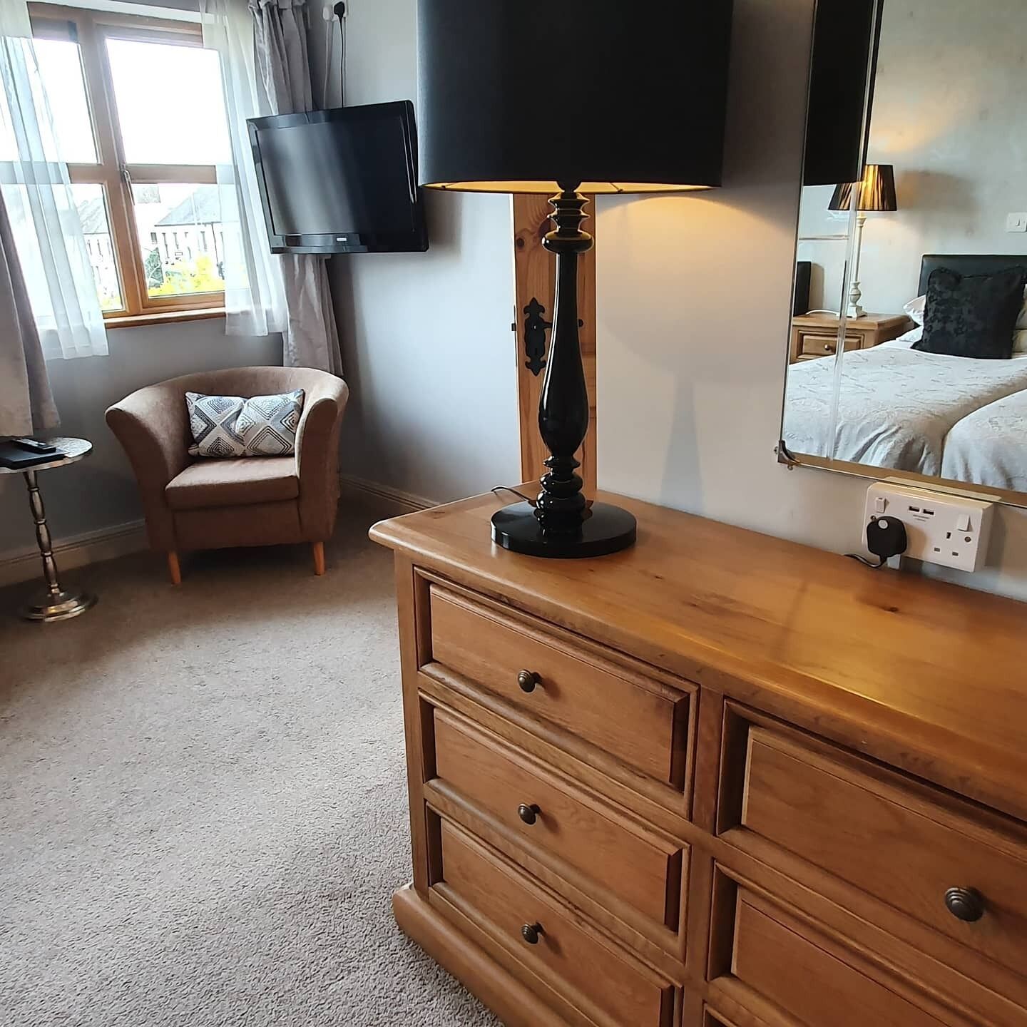 Une pièce comprenant une commode en bois, un téléviseur fixé au mur, une chaise et une table de chevet avec une lampe.