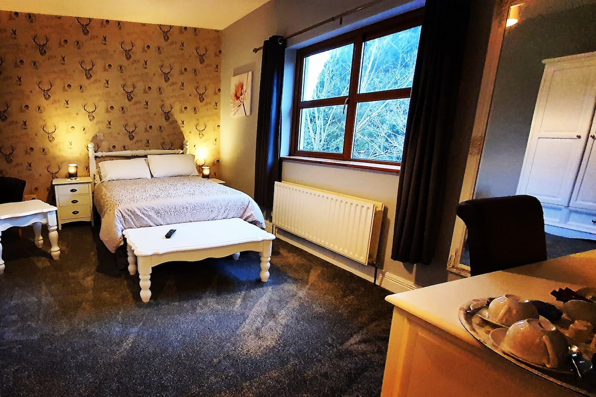 Quarto com cama, mesas de cabeceira, cômoda, radiador e janela com cortinas.