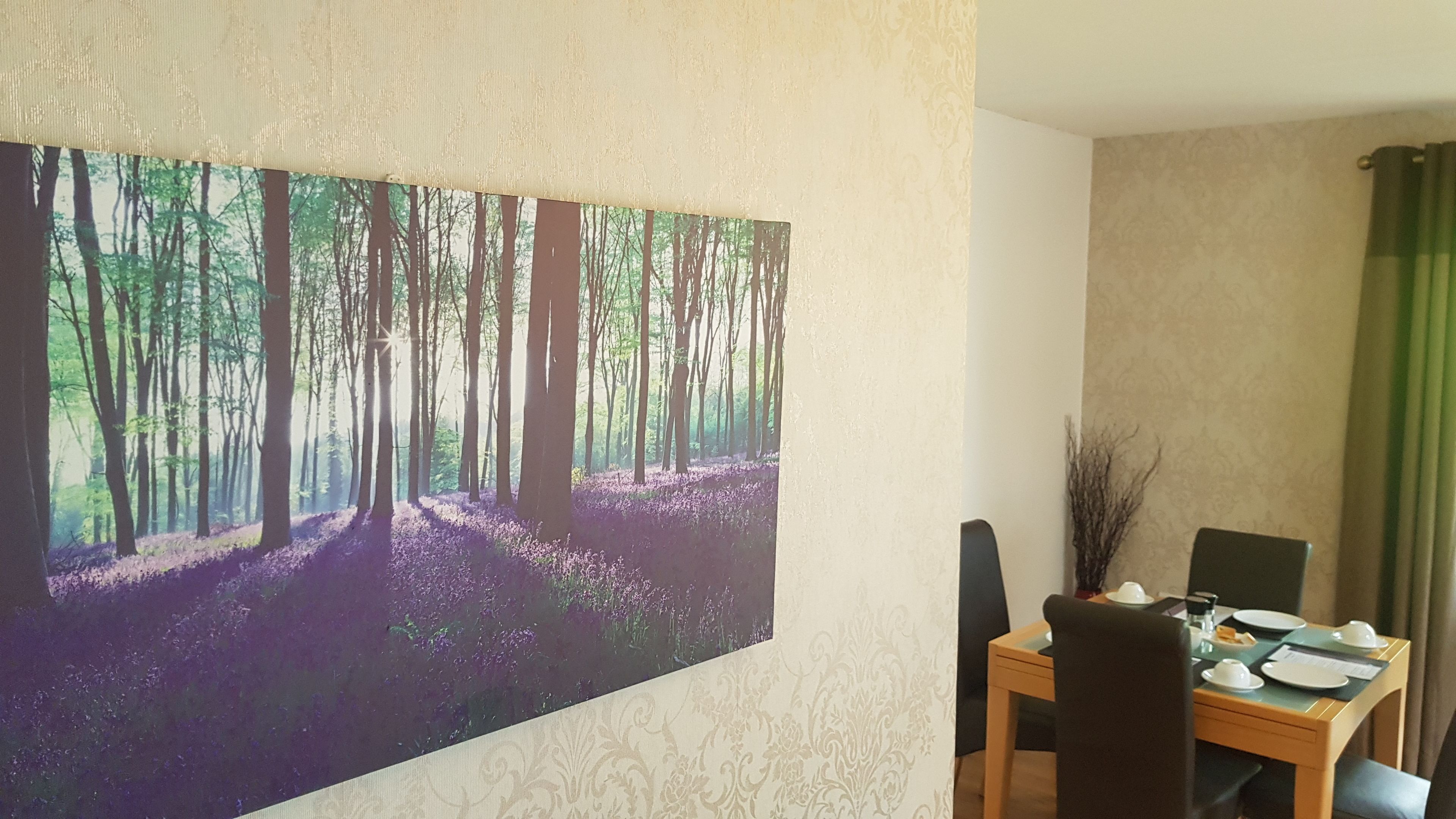 Un comedor con una mesa dispuesta para dos personas, un cuadro de un bosque en la pared y vistas al exterior a través de una ventana.