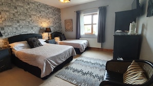 A hotel room with two beds, a nightstand with lamps, a window with curtains, a radiator, a patterned rug, and a chair. - Church View B&B (Gurteen)