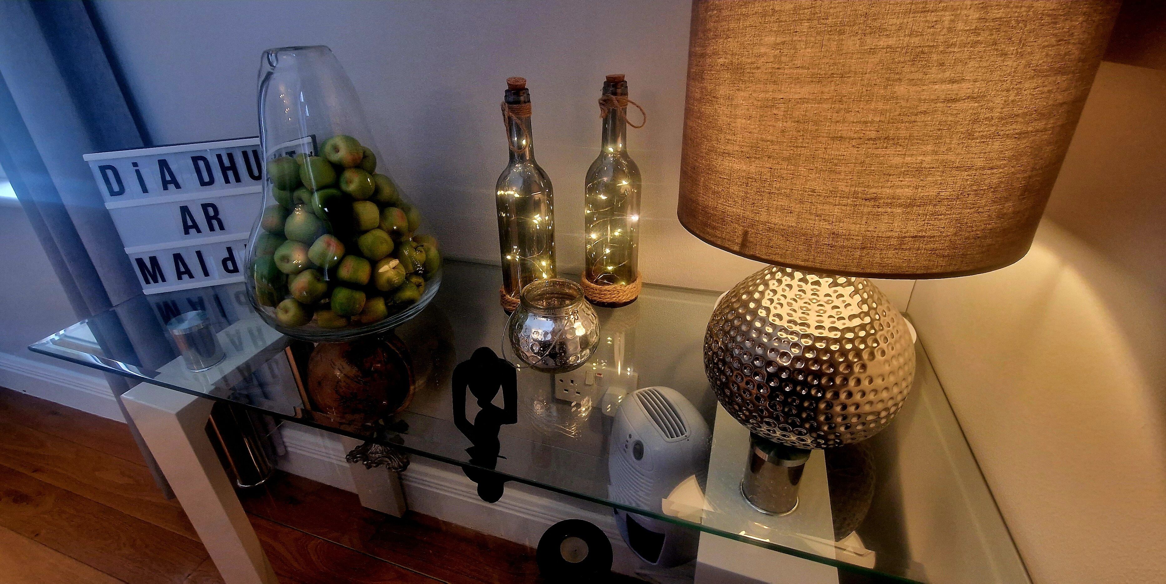 A glass table with decorative items including a jar of apples, two bottles, a lamp, and a sign with text.