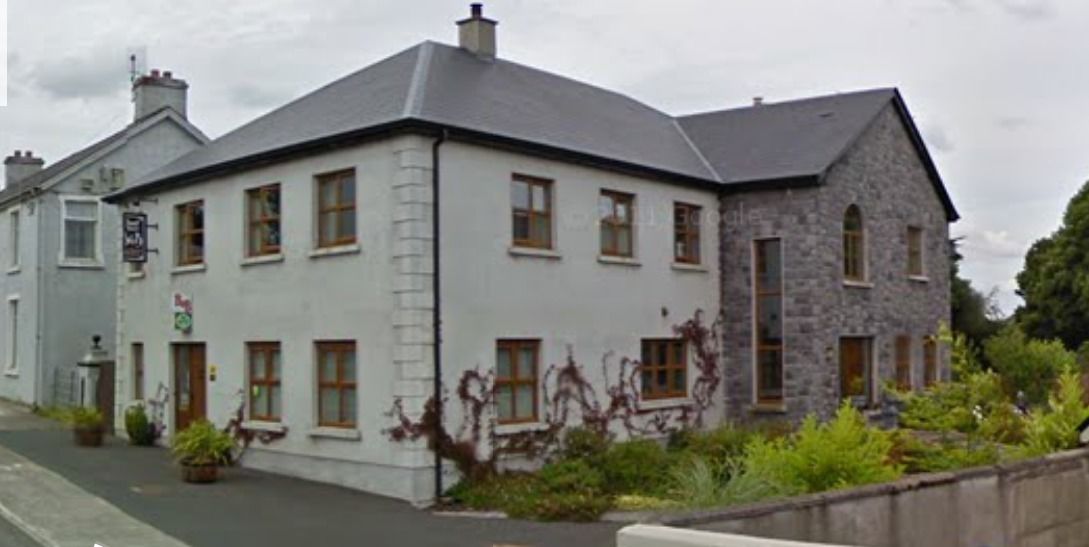 A two-story building with a white facade and stone accents, featuring multiple windows and a small garden area in front.