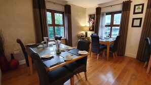A dining area with a wooden table set for four, surrounded by chairs. There are two windows with curtains, a lamp on the table, and a vase with flowers. - Church View B&B (Gurteen)