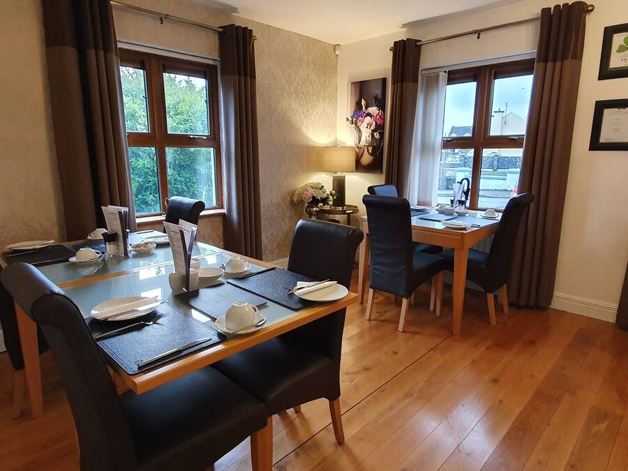 Comedor con una mesa de madera dispuesta para cuatro personas, rodeada de sillas. Hay dos ventanas con cortinas, una lámpara sobre la mesa y un florero con flores.