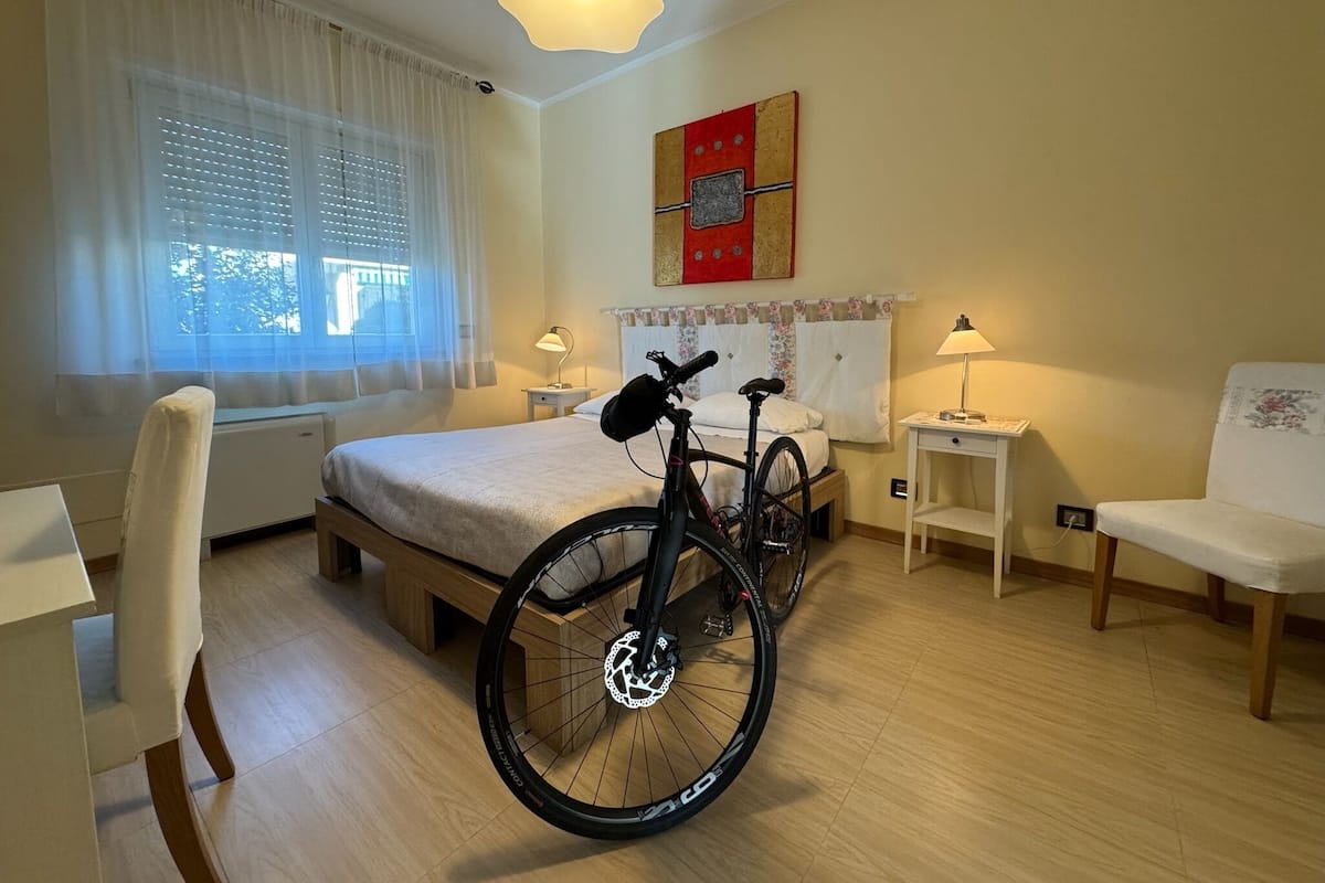 Hotellihuone, jossa on sänky, polkupyörä, tuoli, työpöytä ja ikkuna, jossa on verhot.