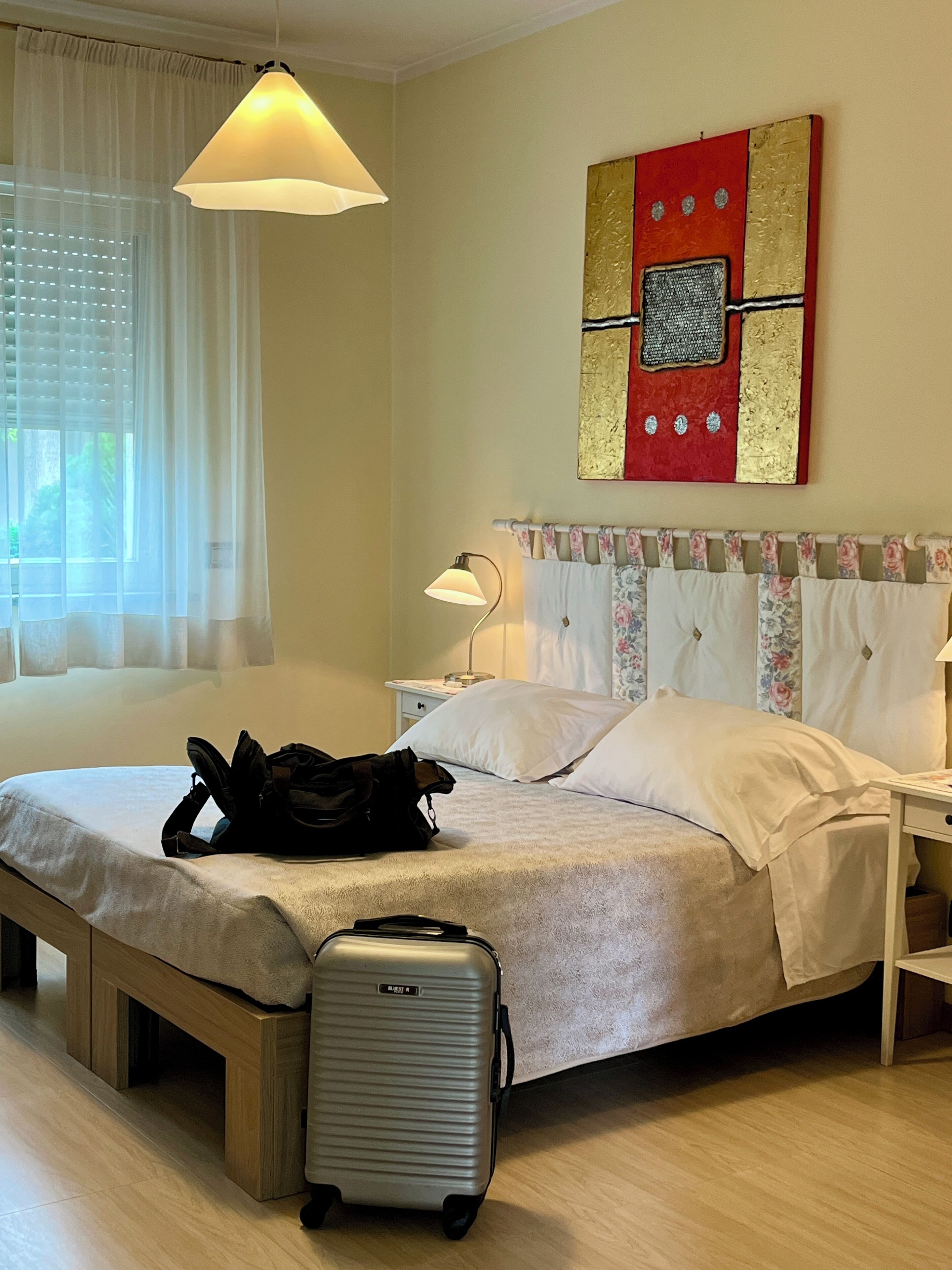A hotel room with a bed, a suitcase, a lamp, and a painting on the wall.